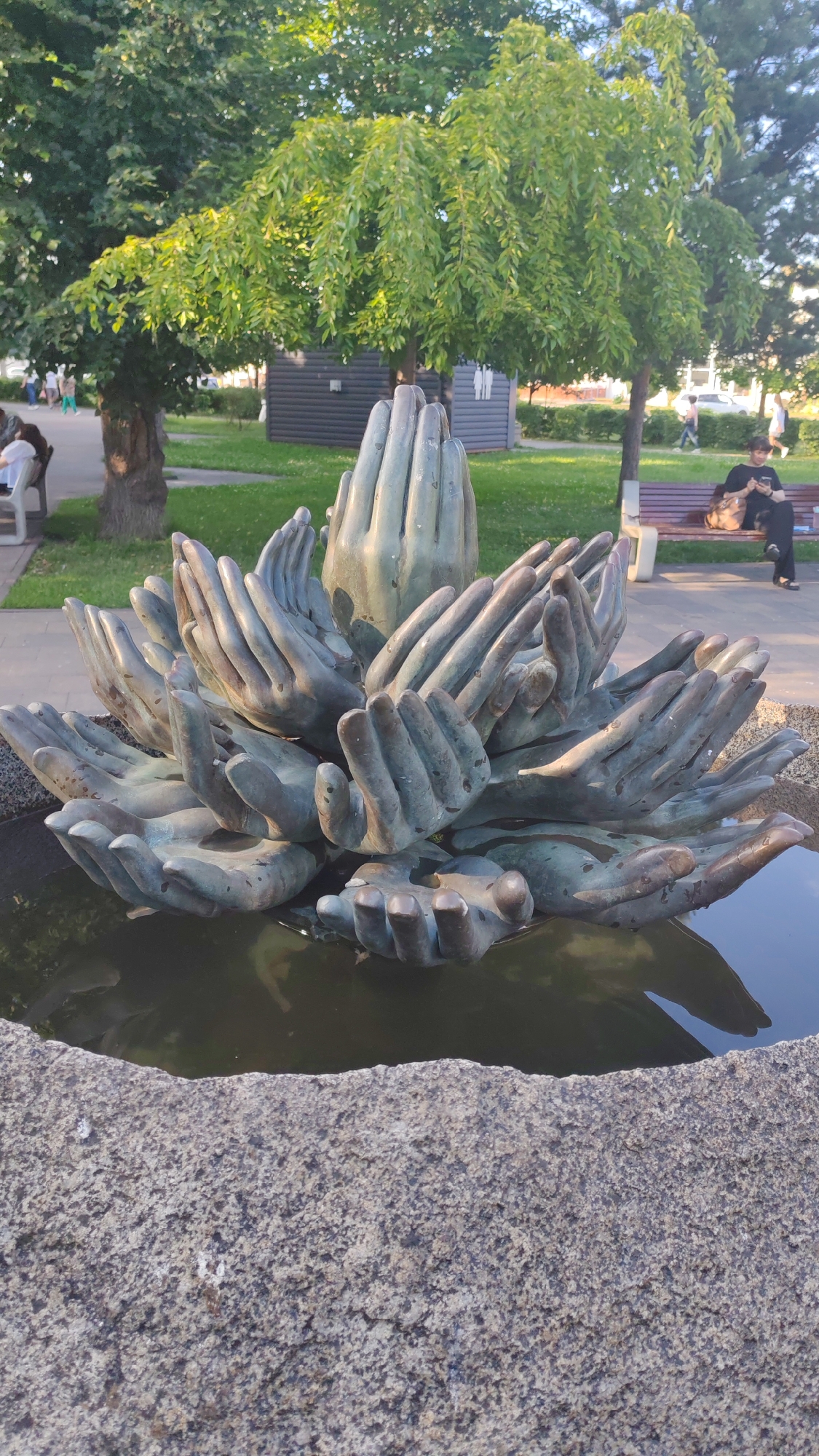 A bronze sculpture of many hands is located in a pond with a stone border in the foreground. The hands are arranged in a circular pattern, with some pointing upwards and others outwards. In the background, there is a grassy area with trees and people walking or sitting on benches. A building with a sign indicating restrooms is also visible. The scene appears to be in a public park setting on a sunny day.