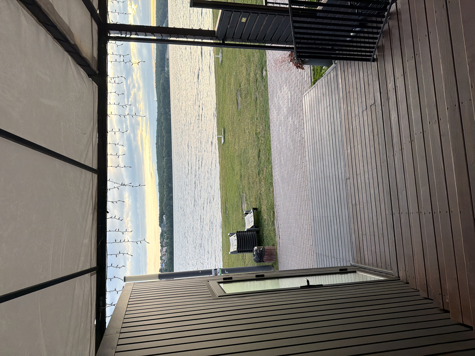 The image depicts a view from a covered wooden patio overlooking a large body of water, likely a lake or river. The patio has a wooden floor and stairs leading down, and a dark wooden wall with a glass door on the left. String lights hang from the white fabric ceiling of the covered area. In the distance, across the water, is a tree-lined shore with buildings visible on a hill. The water is calm with ripples, reflecting the cloudy sky and the late afternoon or early evening light. On the grassy area near the water's edge, there are stacked lounge chairs and a single lounge chair. A trash can with a black bag is also present. The overall setting suggests a recreational area or a waterfront establishment.