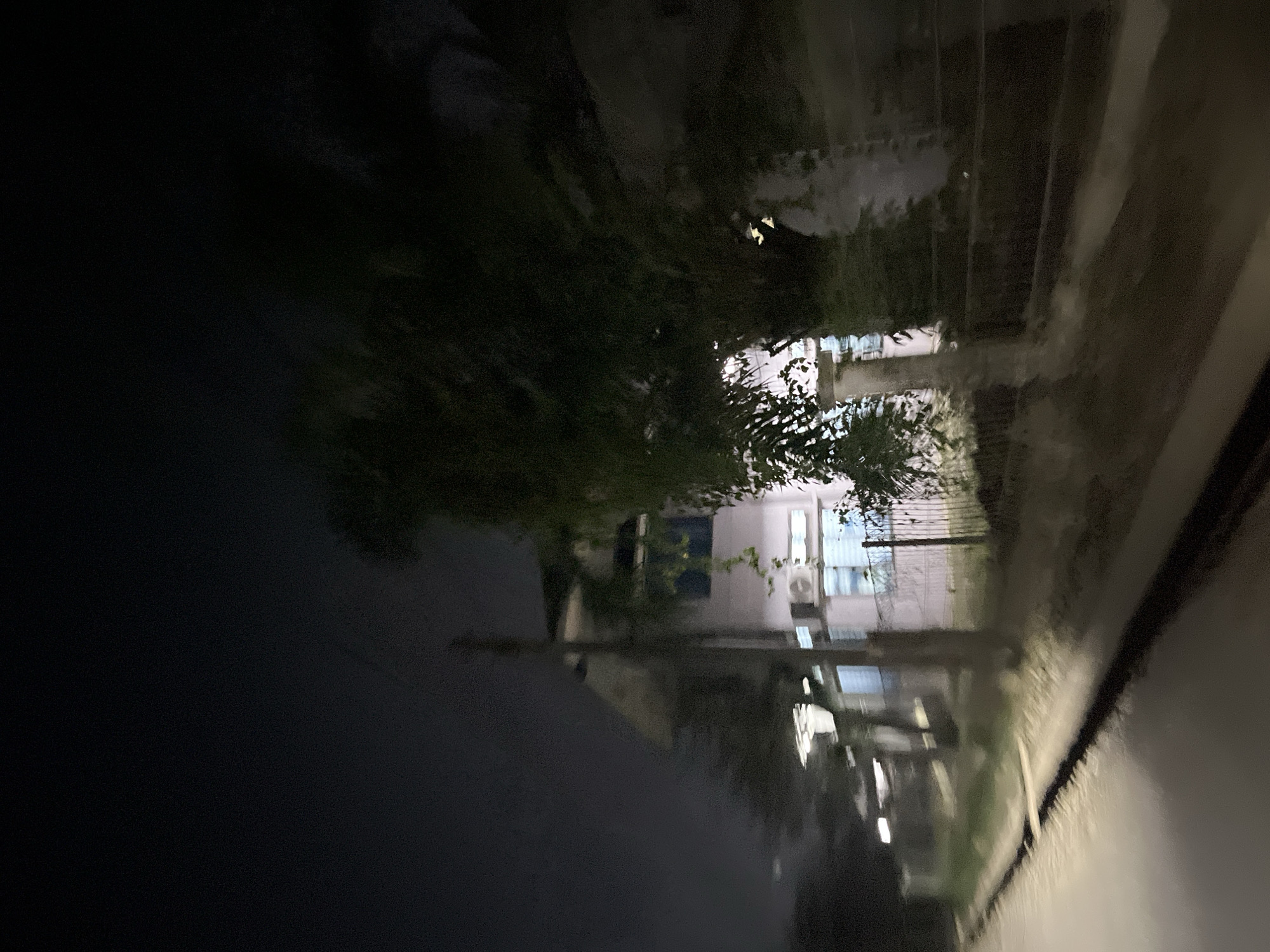 The image depicts a scene at night, possibly in an urban or semi-urban environment in Kesehatanbandar, Indonesia. The primary subject appears to be a building with multiple windows, some of which are illuminated, suggesting it's nighttime and the building is occupied or lit from within. There are also trees and foliage present, casting shadows and adding to the overall atmosphere. The ground or a surface in the foreground is dark and reflective, possibly indicating wet conditions like rain or a puddle, which is further suggested by the hazy and somewhat blurry quality of the image, potentially from moisture on a lens or in the air.
The overall impression is one of a quiet, possibly damp, evening. There are no people or specific objects clearly identifiable beyond the building and vegetation. The lighting is dim, consistent with nighttime. The reflection in the foreground could be of the building and the sky, creating a mirror-like effect. The scene is presented from a low angle, looking up towards the building and the night sky. The focus is soft, giving the image a dreamlike or impressionistic quality. No text is visible in the image.