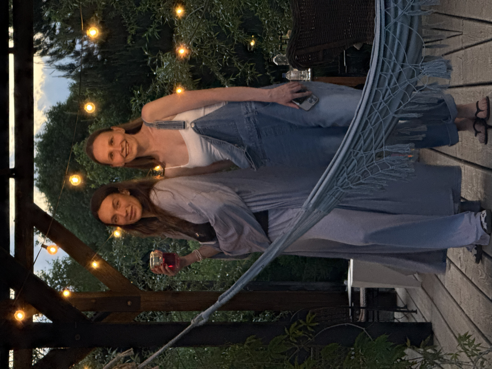 Two women are posing together on a wooden deck, likely outdoors. The first woman is wearing a white tank top and denim overalls, holding a cell phone in her hand. She has long hair and is smiling. The second woman is wearing a long-sleeved, light blue shirt or jacket and is holding a glass filled with a red beverage. She also has long hair and is smiling. A light blue hammock is visible in the background. Strings of lights are strung across the deck and into the trees, suggesting an evening setting. The surroundings include greenery and parts of a wooden structure or fence. There's nothing suggesting the city of Dobroe, Russia.