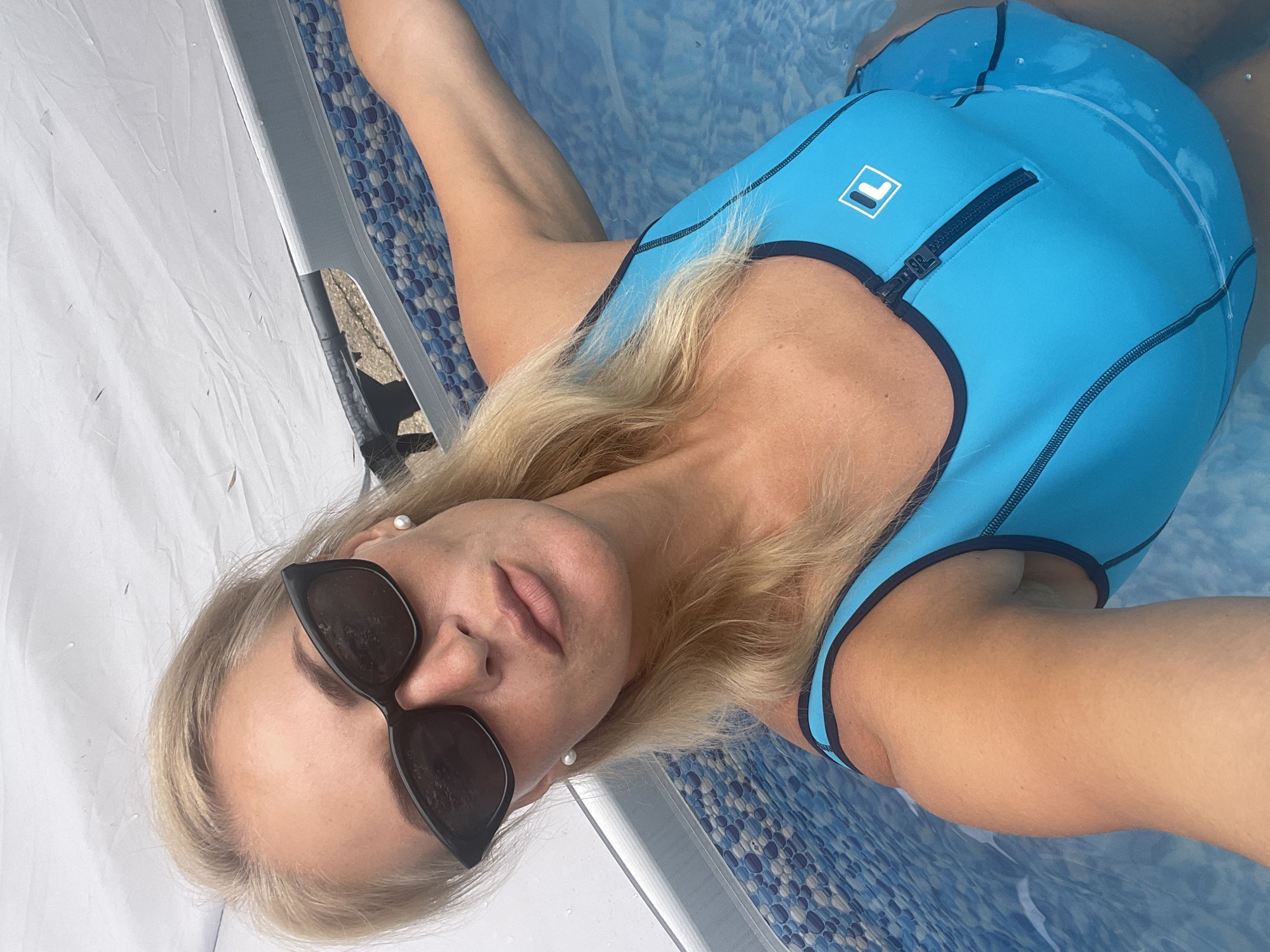 A blonde woman wearing sunglasses and a blue swimsuit is taking a selfie in an inflatable pool. The pool has a patterned liner and is partially covered by a white tarp. The woman has pearl earrings and her hair is wet. The image appears to be taken outdoors. The location is specified as Baranovka, Russia.