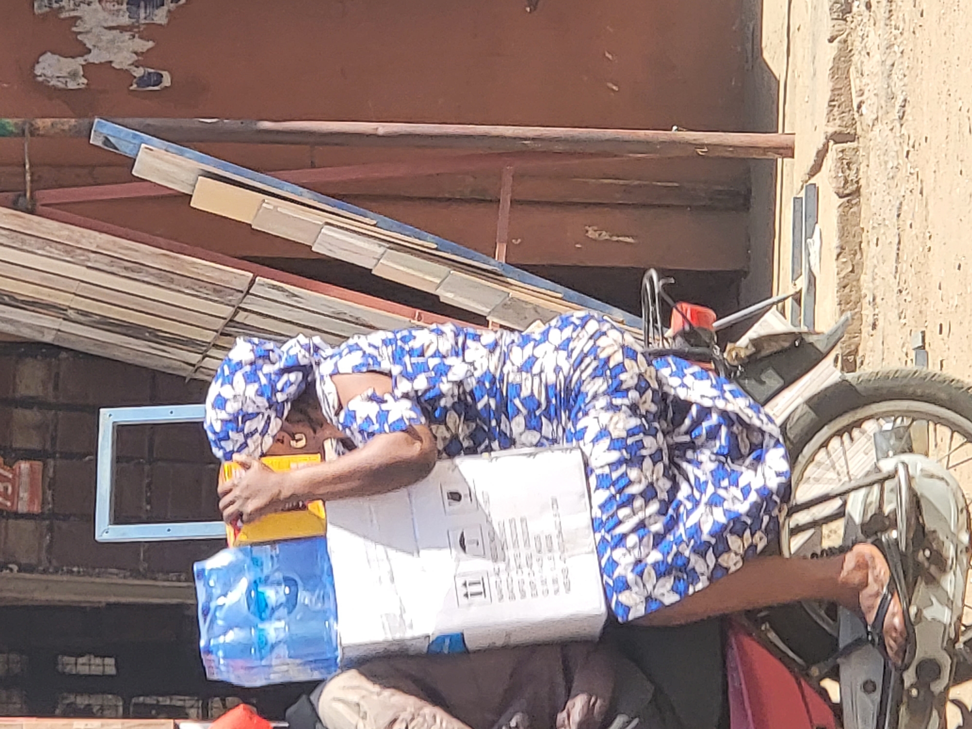 In Maiburiji, Nigeria, a woman in a blue and white floral patterned dress and headwrap sits on a red and black motorcycle, holding a stack of items against her body. These items include a case of bottled water, a yellow box, and a white cardboard box with printed symbols and text. Her bare right foot rests on the motorcycle's footrest, clad in a sandal. The background includes a brick wall, a window frame, and a structure composed of wooden planks and metal beams. A building with a textured exterior is visible to the right.