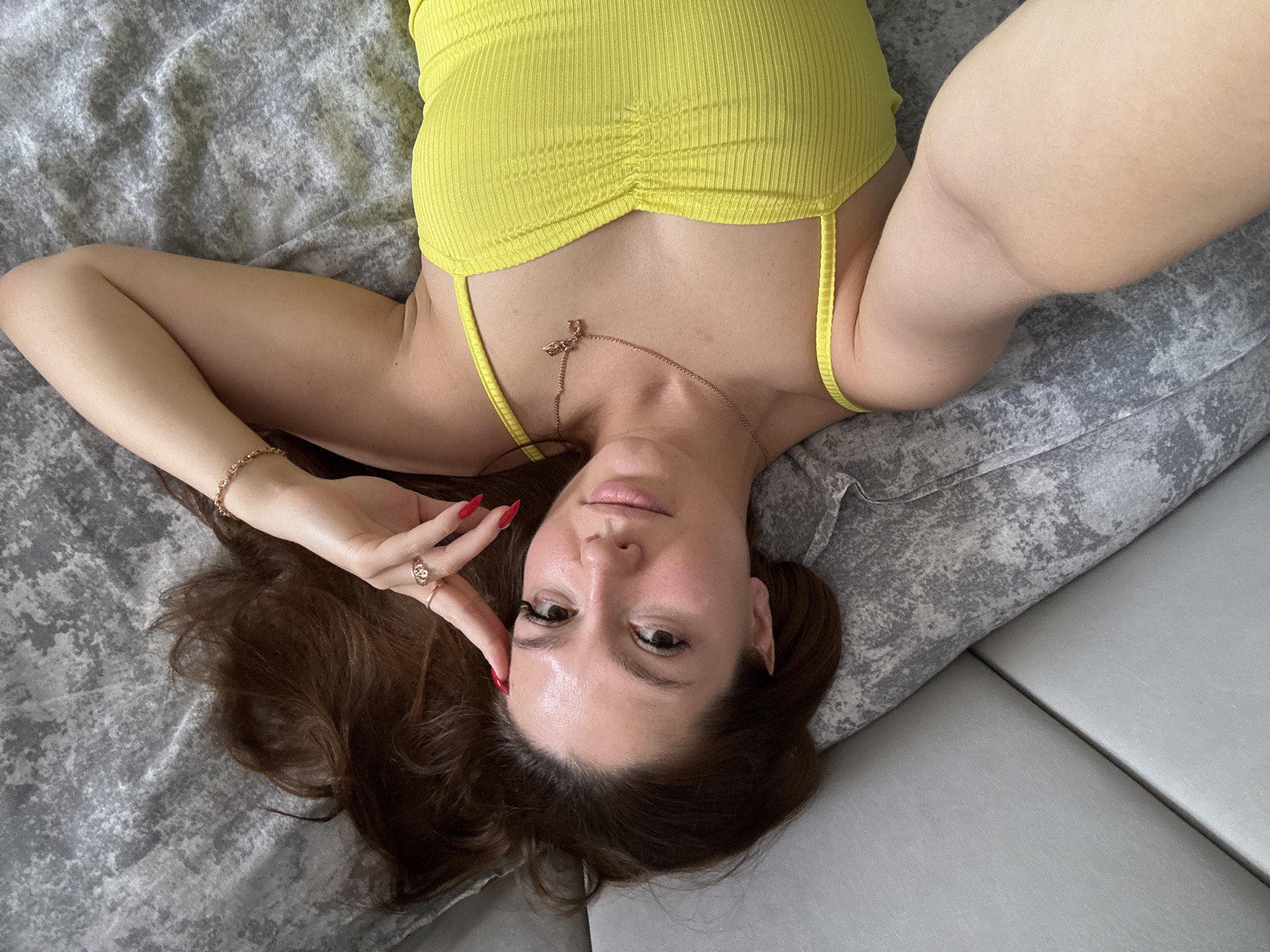 A young woman with long, dark brown hair lies on her back on a bed with a gray, textured comforter and a light gray upholstered headboard. She is looking up and to her right, with her left hand resting on her forehead and her right hand resting near her chin. She is wearing a bright yellow, ribbed tank top with thin spaghetti straps. A delicate gold necklace with a small pendant is visible around her neck. She has on rings on her right index finger and right ring finger, and a bracelet on her right wrist. Her fingernails are painted red. The overall lighting suggests an indoor setting, likely a bedroom.