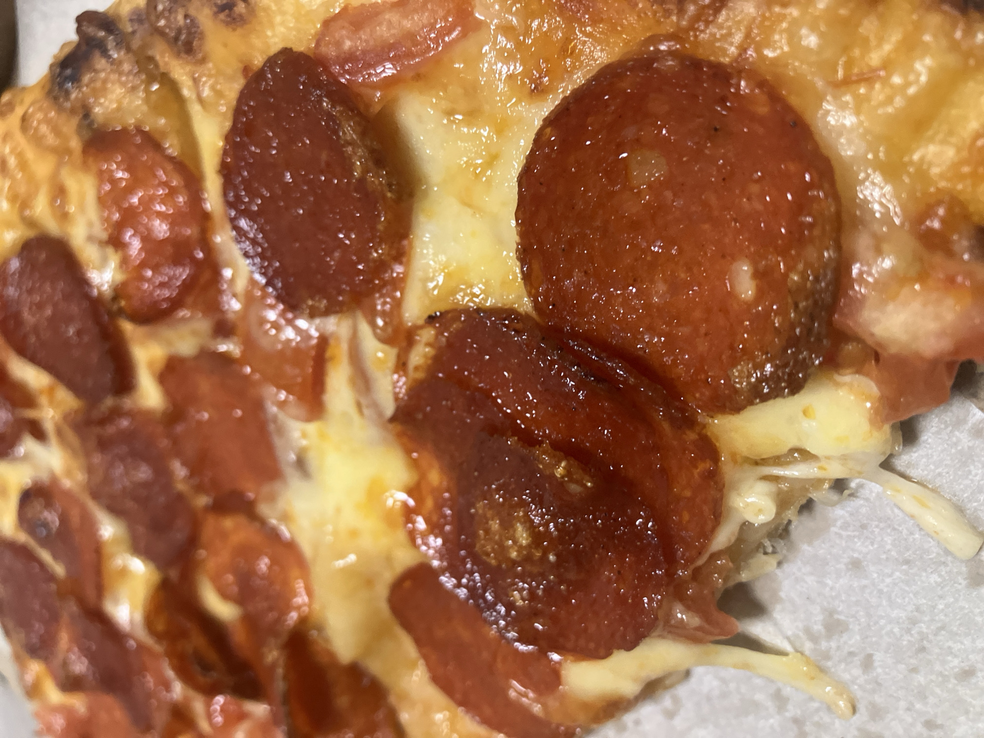 This is a close-up, high-angle shot of a slice of pepperoni pizza. The main subjects are the pizza toppings: numerous round slices of pepperoni and generous amounts of melted cheese.
The pepperoni pieces are reddish-brown, some with slightly curled and crisped edges, and a glossy, almost greasy sheen, indicating they are hot and have rendered some fat. Small bubbles or texture variations are visible on their surface. Beneath and between the pepperoni is a thick layer of melted cheese, predominantly a creamy white and golden-yellow color, with some areas appearing more browned or bubbly from baking. At the bottom right of the image, the cheese is visibly stretched and stringy, suggesting the slice has just been lifted or pulled apart.
The pizza crust is visible along the top and left edges, showing a golden-brown color with some darker, slightly charred spots, indicating a well-baked quality. The texture of the crust appears somewhat irregular, hinting at an airy or bubbly structure. The overall impression is that of a warm, appetizing pizza.
There is no specific activity depicted other than the implicit action of the pizza being presented or consumed. The notable details include the glossy pepperoni and the stretchy, melted cheese, suggesting the pizza is fresh and hot. There are no people visible, and no visible text in the image. The image itself does not provide any specific visual cues to confirm the location as Chaikovsky, Russia. The setting appears to be an indistinct background, possibly a pizza box or simple plate.