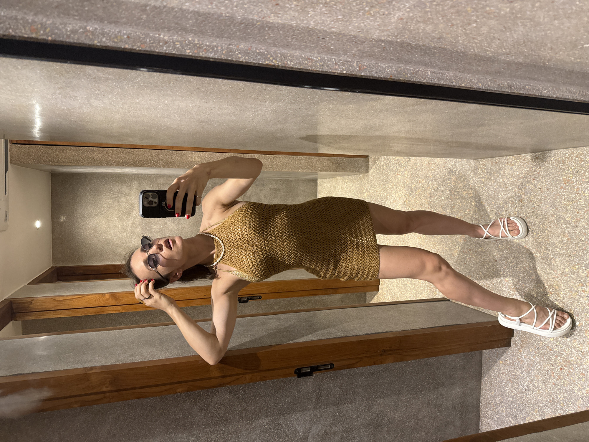A woman stands in front of a mirror, taking a selfie. She is wearing a gold, knit halter dress, white strap sandals, and sunglasses. She has on a pearl necklace and is touching her hair. The setting appears to be a dressing room or bathroom, with wooden doors and textured walls.