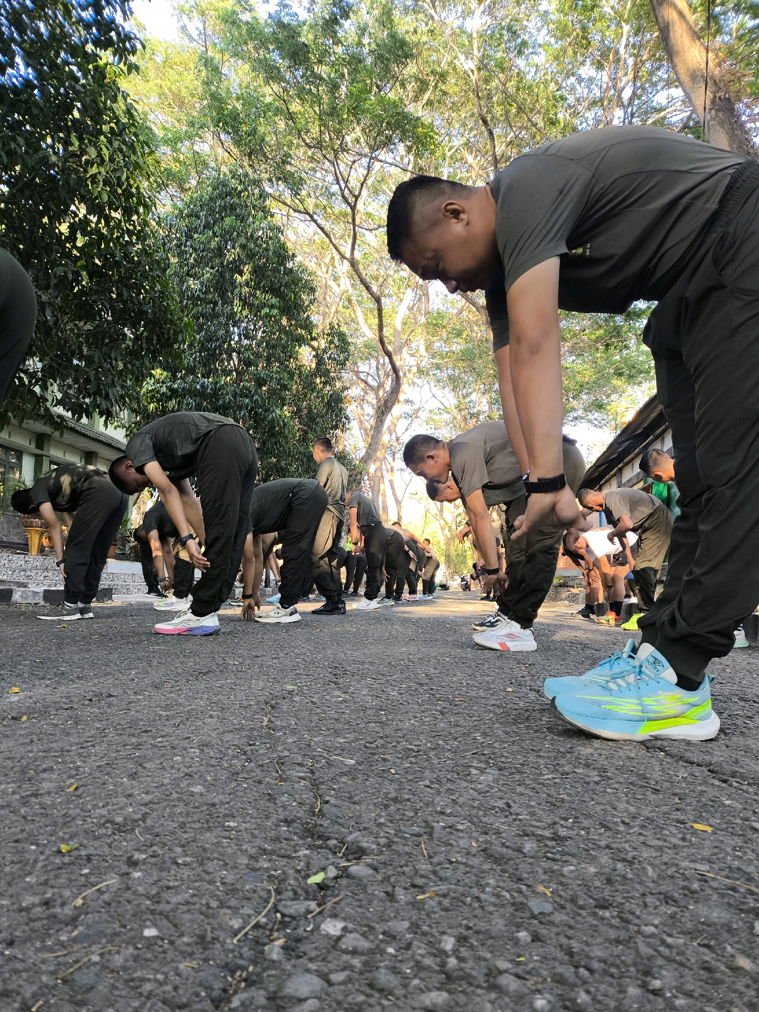 In an outdoor setting in Unknown, Indonesia, a group of men in dark green athletic uniforms are performing stretches on an asphalt surface. The men are positioned in a line, with their backs to the camera in the foreground and midground, and their faces mostly obscured as they bend at the waist. The environment features lush green trees with dappled sunlight filtering through, suggesting a park or an open area adjacent to buildings. A building with a light-colored facade and dark roofline is visible in the background, hinting at a training facility or institutional grounds. The asphalt is textured with pebbles and small debris, with some scattered leaves. The overall scene depicts a synchronized physical training exercise.