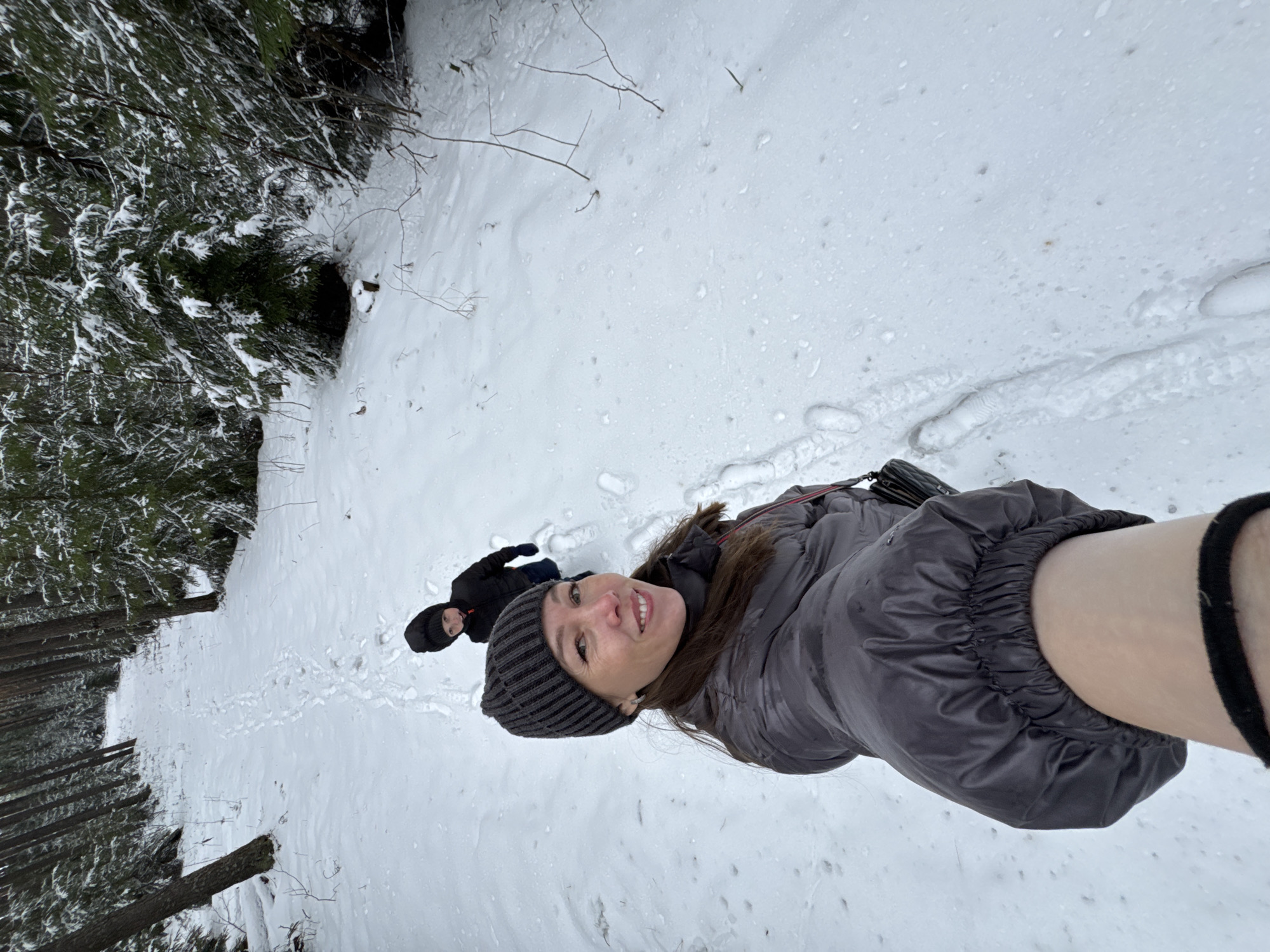 A selfie photograph depicts a woman and child in a snowy environment. The woman, wearing a gray puffer jacket and a gray striped beanie, smiles towards the camera. A child, dressed in dark clothing, stands a short distance behind her. The ground is covered in snow, with visible footprints leading away from the foreground. To the left, a dense forest is visible, characterized by snow-covered trees. This scene likely takes place in the city of Chaikovsky, Russia.