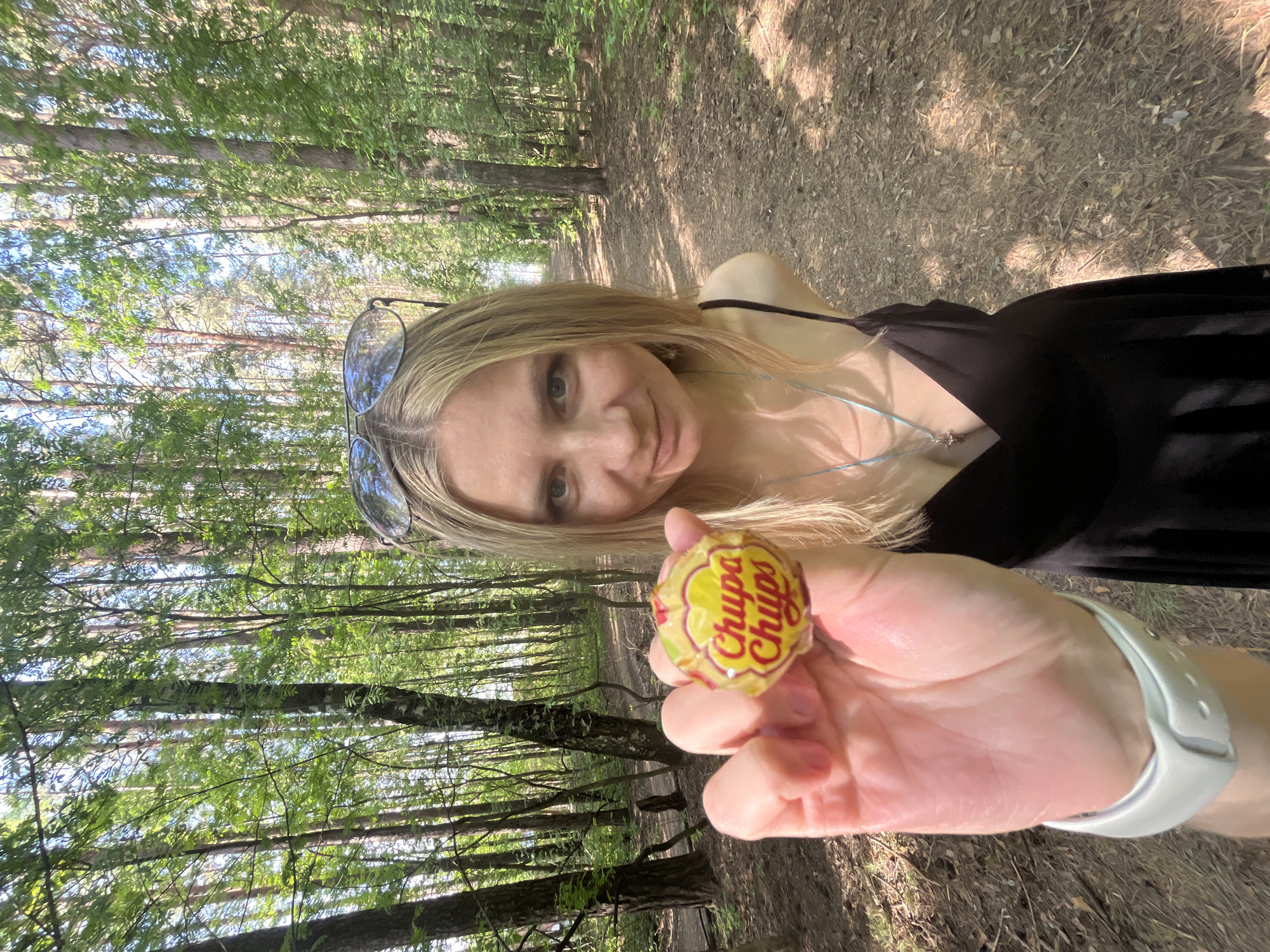 A woman with blonde hair and sunglasses poses in a forest holding a Chupa Chups lollipop. She wears a black dress and a white smartwatch. The location appears to be in a wooded area near Chaikovsky, Russia, based on visual characteristics.