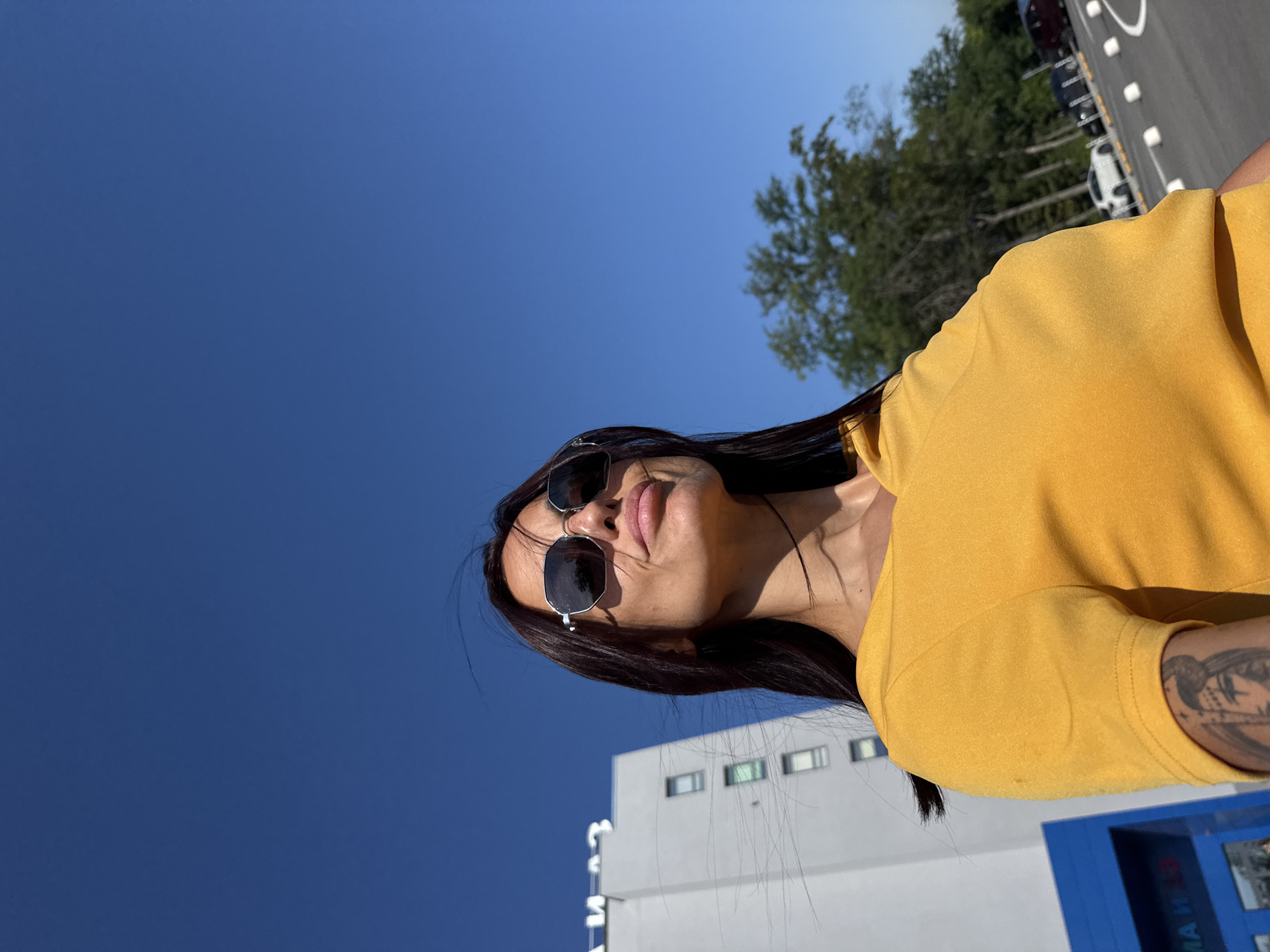 The image captures a woman with long, dark hair and sunglasses, wearing a yellow t-shirt, standing outdoors under a clear blue sky. In the background, a tall, light-colored building with windows is visible, along with trees and a paved area with white bollards and some parked cars. The building appears to be branded with Cyrillic lettering. Given the visible Cyrillic text on the building, the location is likely the city of Pleven, Bulgaria, potentially near a business or administrative center. The lighting suggests it is daytime. The woman is looking upwards and to her right, with her lips slightly pursed. A tattoo is visible on her left arm.
