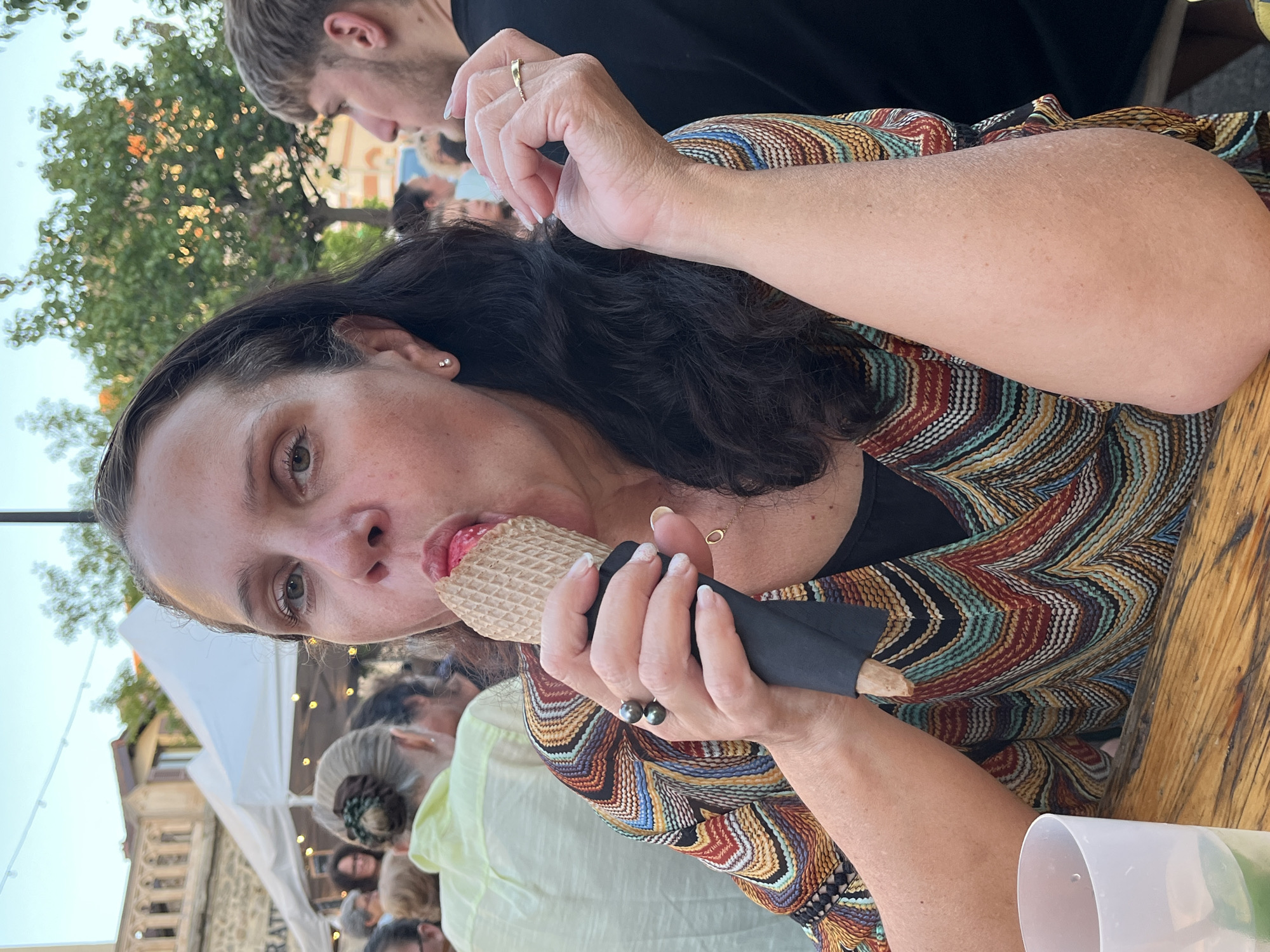 A woman with dark brown hair is eating an ice cream cone at an outdoor event. She is wearing a colorful, geometric-patterned top. She is holding the cone in her right hand and has an ice cream dripping slightly onto her fingers. Her left hand is resting on her chest. In the background, there are tents, trees, and other people. The scene appears to be in a town square or market. The setting suggests an informal gathering or festival.