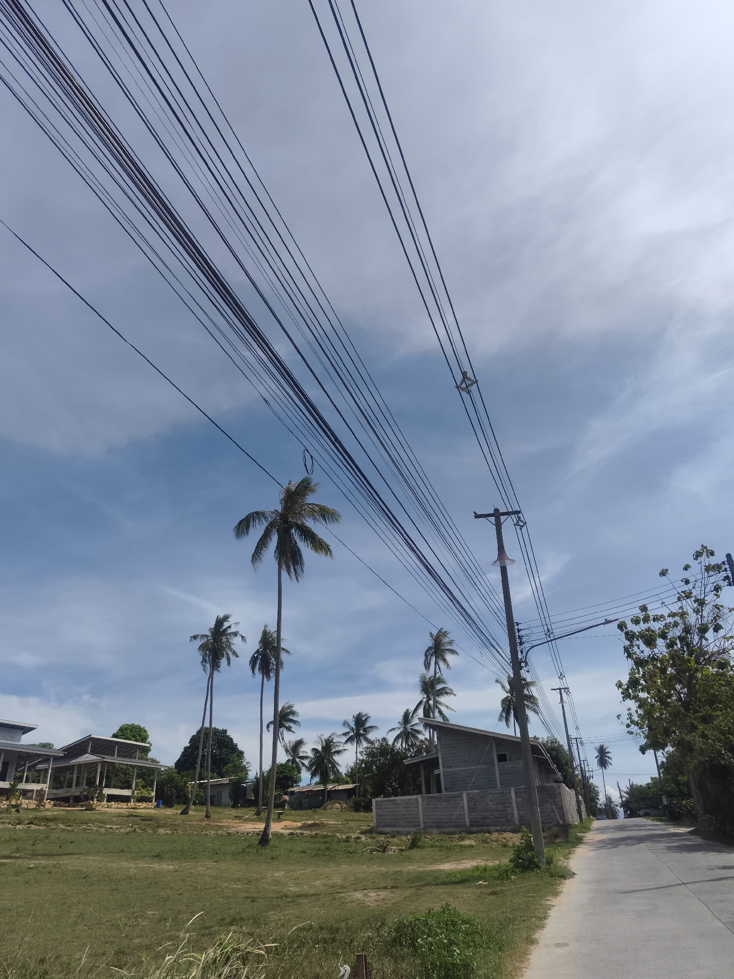 The image shows a street scene in Baan Tai, Thailand. A paved road extends into the distance. Utility poles line the road, with numerous electrical wires stretching across the sky. Buildings under construction are visible on either side of the road. Palm trees are scattered throughout the landscape. The sky is blue with scattered clouds. A grassy area is present on the left side of the road. Vegetation includes trees and shrubs.