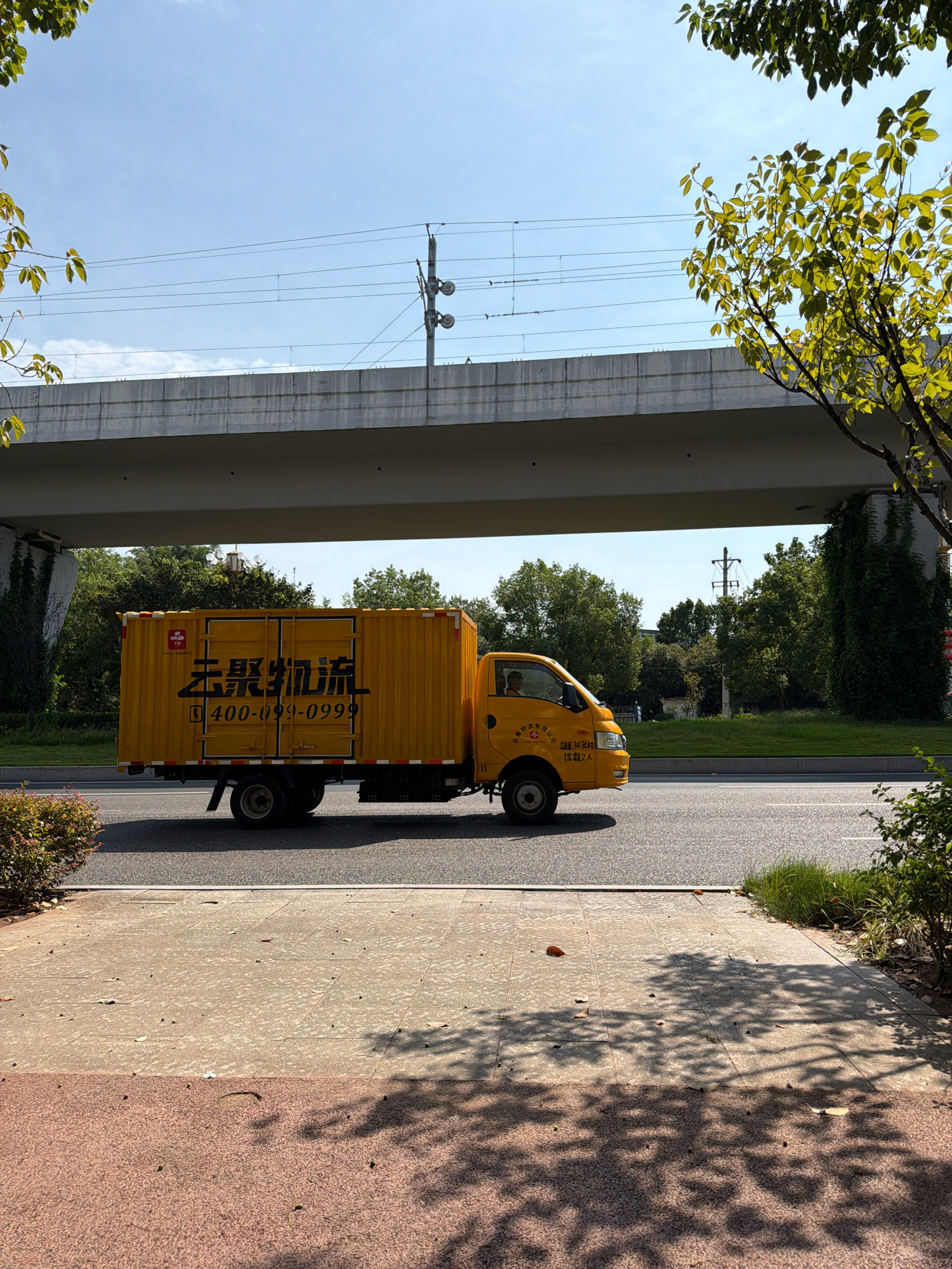 A yellow cargo truck with "Jingyu Logistics" written on the side is driving on a road under an elevated railway overpass. The truck is a two-axle vehicle with a closed cargo container. The side of the container has large black Chinese characters and a phone number: 400-099-0999. The truck is moving from left to right. In the foreground, there is a sidewalk made of concrete tiles, with some fallen leaves. In the background, there are trees and greenery. The sky is blue with some white clouds. The scene appears to be in an urban or suburban environment.