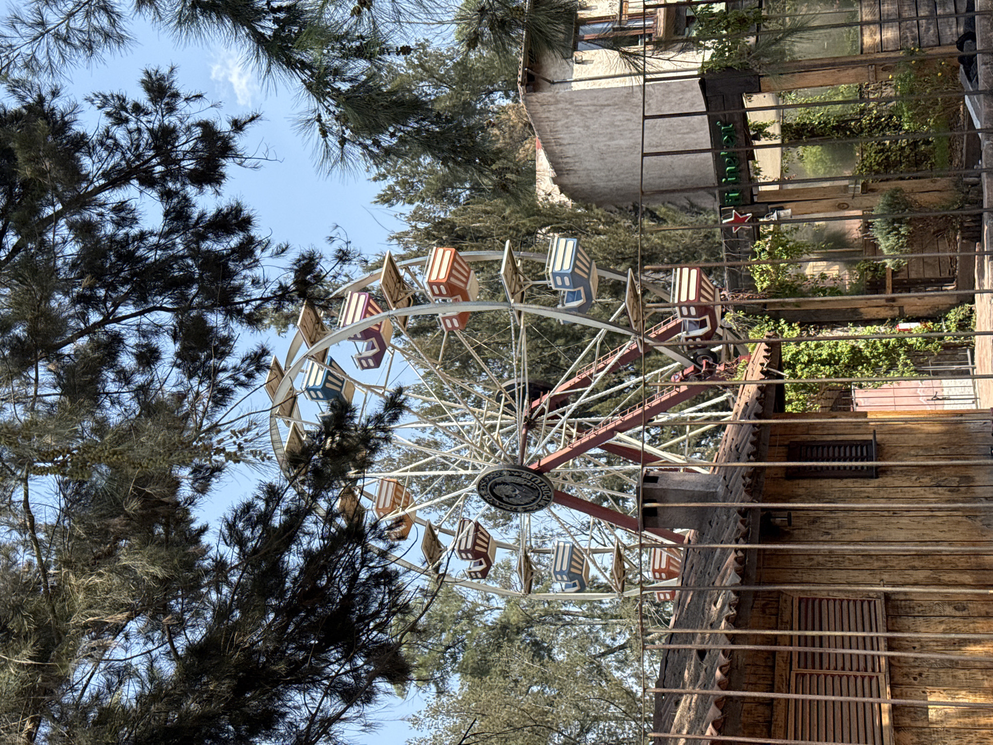 The image shows a Ferris wheel in Domingo Arenas, Mexico. The Ferris wheel has red and white supports and a white wheel with gondolas in alternating colors of red, white, and blue. It is surrounded by trees, including pine trees on the left. A building with a wooden facade and barred windows is in the foreground. Behind the Ferris wheel, there is another building with a sign that includes a star. The sky is visible through the trees, and appears blue.