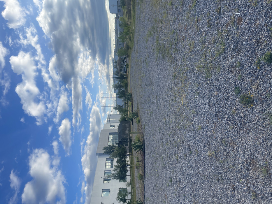 This image captures an industrial or commercial park setting in Unknown, Unknown, under a partly cloudy blue sky. The foreground is dominated by a large, unpaved lot covered in grey gravel, with sparse green weeds and small yellow wildflowers growing between the stones. In the midground, a landscaped median features several agave plants and small deciduous trees. Directly behind this, a white, modern two-story building with multiple reflective windows and a grey-paneled entrance area is visible on the left. A dark, netted archway structure extends from the building towards the landscaped area. To the right and further in the background, additional industrial buildings with lighter-colored roofs and multiple tall poles (possibly flagpoles or light poles) are present. A few parked vehicles are discernible in the distance near these structures. No people are visible, and no specific actions or interactions are occurring. The environment appears clean and well-maintained, suggesting a business or manufacturing campus.