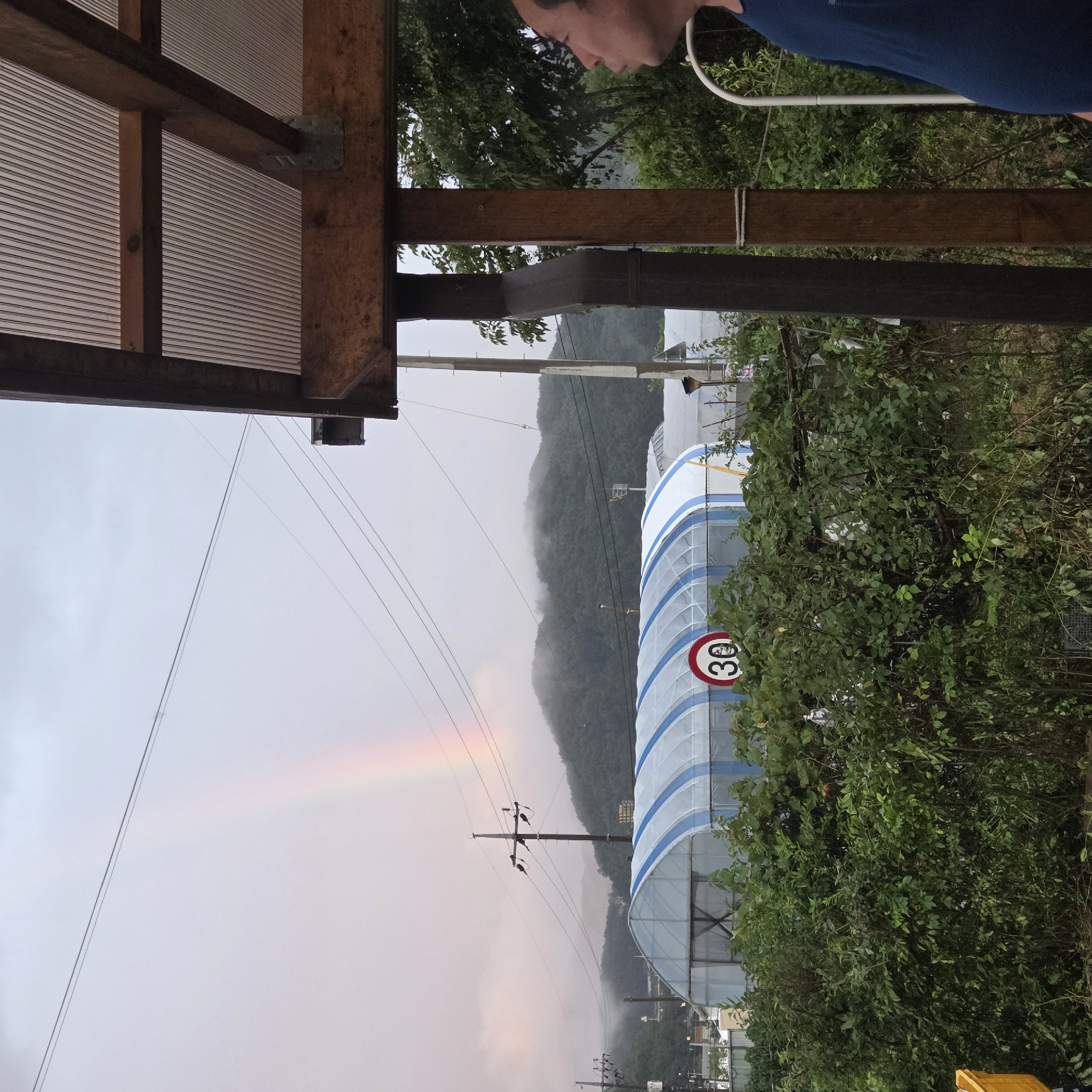 In Dunnae-myeon, South Korea, under a cloudy sky, a faint rainbow arches across the horizon, partially obscured by mist that blankets distant, tree-covered hills. In the foreground, a large, greenhouse-like structure with blue and white striped roofing is visible, surrounded by dense green foliage. A circular speed limit sign with the number "30" is mounted on a post near the greenhouse. The scene is viewed from under a wooden overhang with a corrugated plastic roof. On the right side of the image, a man in a blue shirt is partially visible, looking towards the landscape. Electrical poles and wires crisscross the sky, indicating an outdoor, likely rural or semi-rural, setting.