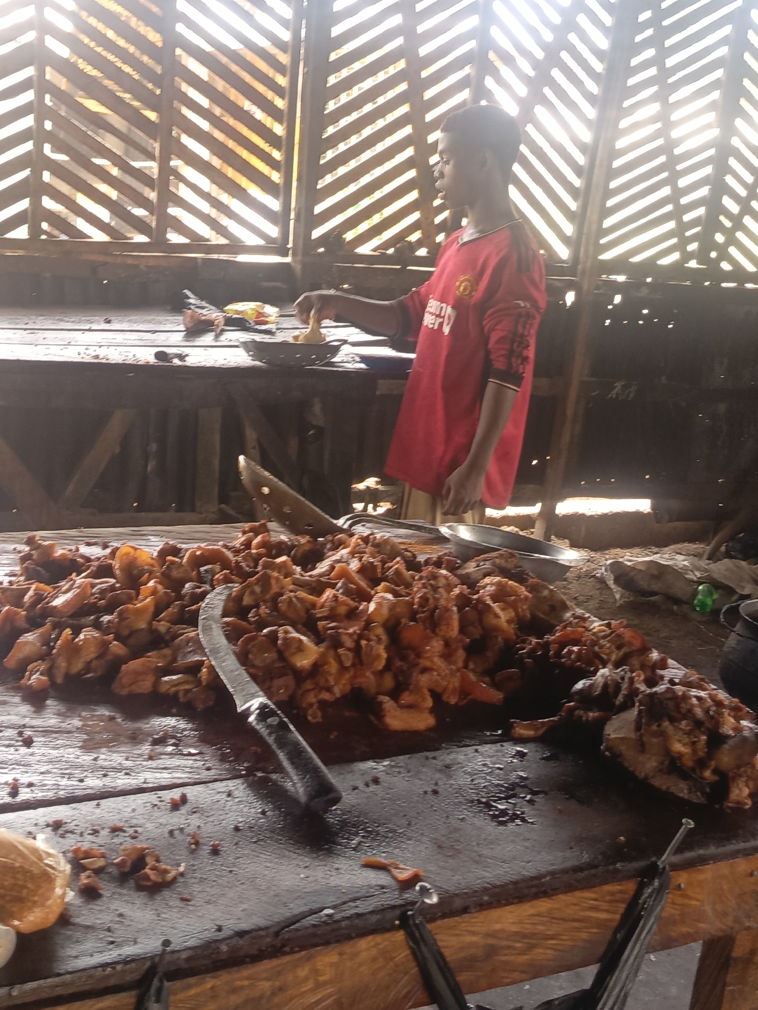 A person wearing a red Manchester United T-shirt stands behind a table laden with fried food. The food appears to be meat, possibly chicken or pork, chopped into irregular pieces. A large knife with a black handle lies among the food. The person is using a slotted spoon or similar utensil to transfer some of the food into a metal bowl or dish on another part of the table. The setting appears to be an indoor or covered area, with sunlight streaming through a lattice-style window or wall in the background. The construction seems rough and rustic, suggesting an informal eatery or roadside stand in the city of Unknown, Unknown.