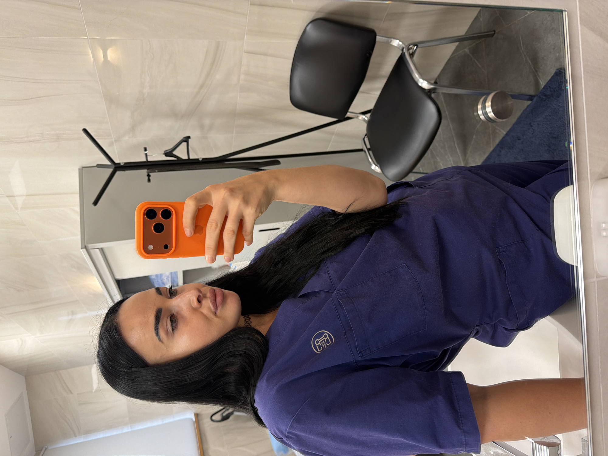 A mirror selfie captures a woman indoors, likely in Saint Petersburg, Russia. The woman, with long dark hair, a calm expression, and subtle makeup, is dressed in a dark blue uniform or scrubs, which features a circular white emblem on the left chest pocket. She holds an orange-cased smartphone, equipped with a triple camera system, to take the reflection photo.
The background, visible in the mirror, reveals a clean and modern interior. The walls are adorned with light-colored, possibly marble-patterned tiles, while the flooring reflected in areas with the chairs appears to be a darker grey tile. Two black office-style chairs with shiny chrome frames are positioned in the room. A black stand or rack is also visible. A blue cloth or towel is present on a surface or the floor near the mirror's edge. The setting, characterized by its tiled surfaces and functional furniture, suggests a professional, possibly medical or clinical, environment.