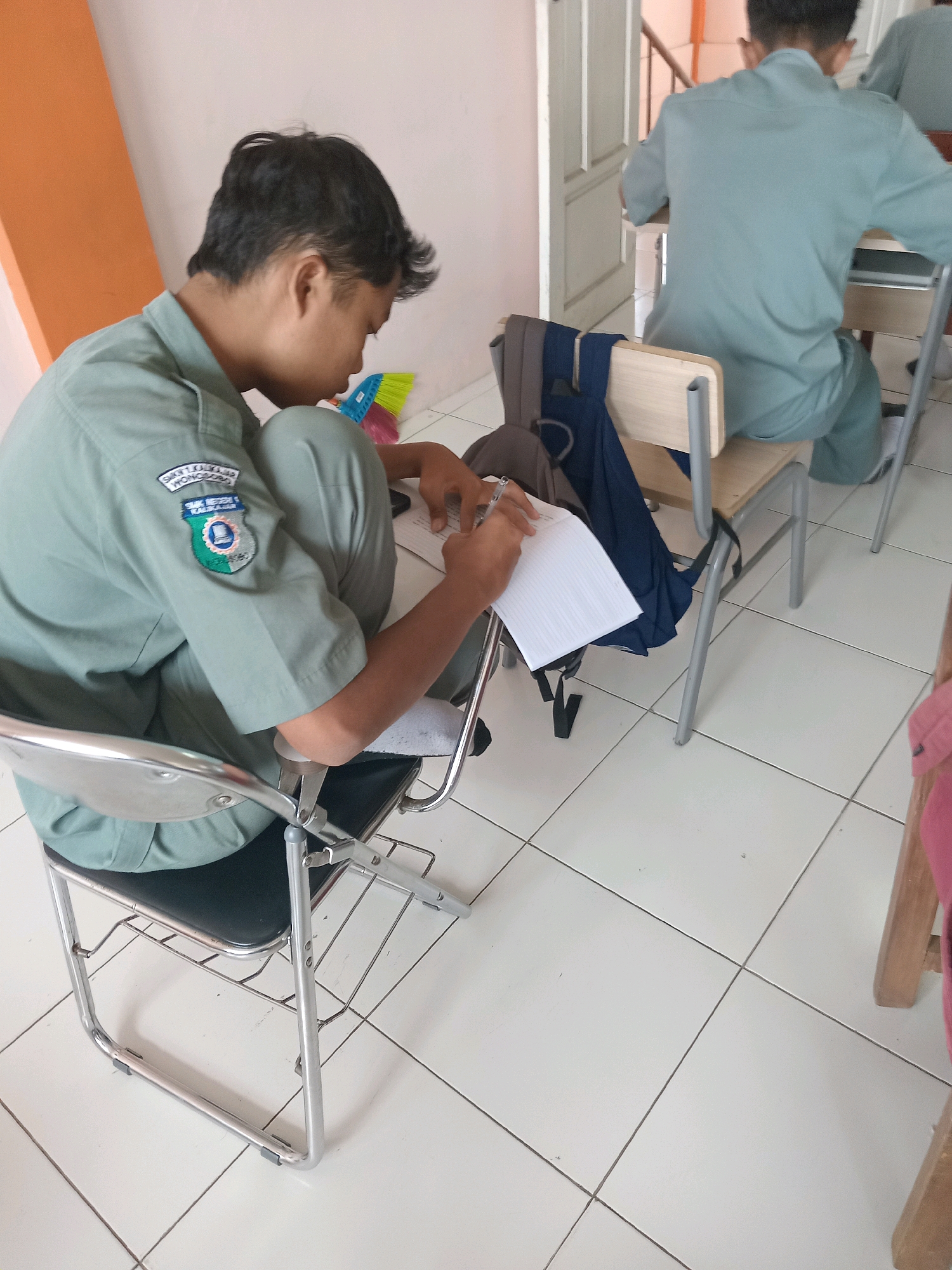 An indoor scene depicts two young males in light green uniforms, likely students, seated at desks in what appears to be a classroom or examination hall. The primary focus is on a young male in the foreground, sitting on a metal-framed chair with a black seat, hunched over a piece of paper and writing with a pen. He has dark hair and is wearing a light green collared shirt with a patch on the left breast featuring a blue and green emblem. His left leg is crossed over his right. Behind him, a dark blue backpack and a grey jacket are draped over his chair. To the right, a second young male, also in a light green uniform, is seated at another desk, facing away from the camera, seemingly engaged in writing as well. The environment is characterized by a white tiled floor and an orange wall on the left side. The presence of desks and the activity of writing suggest an academic setting. The location is identified as Kalikajar, Indonesia.