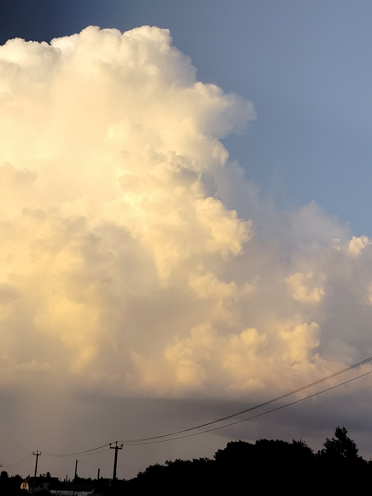 The image depicts a dramatic sky over Yashkino, Russia, dominated by a large, illuminated cumulus cloud formation. The cloud is tinged with warm yellow and orange hues, suggesting the light of either sunrise or sunset. A darker, stormier cloud bank lies beneath it, casting a shadow over the landscape below. Silhouetted against the sky are trees, power lines, and what appears to be a small building or structure. The foreground is dark, with the details of the ground obscured by shadow. The overall impression is one of a powerful natural event unfolding above a quiet, rural setting.