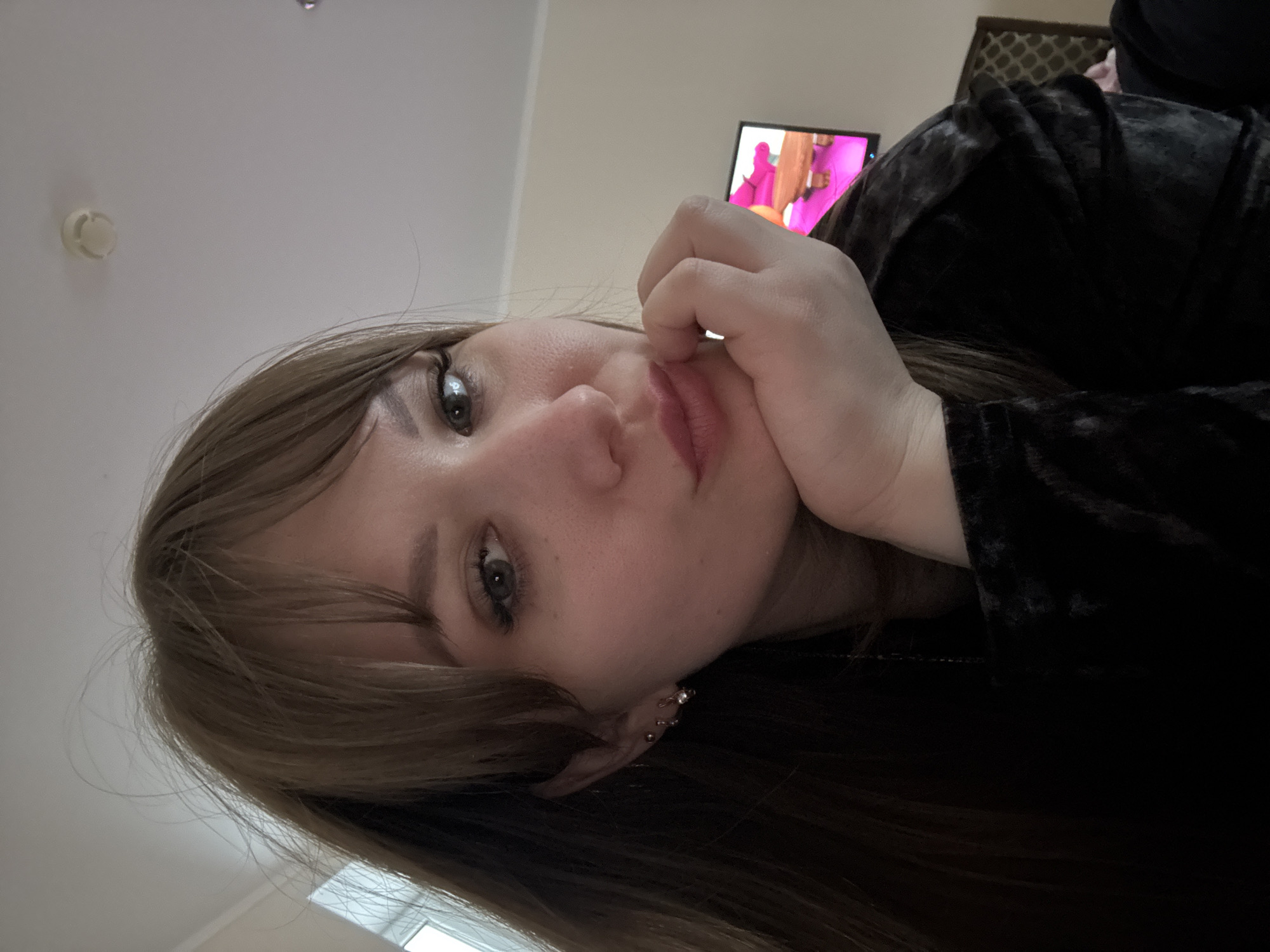 A young woman with light brown hair and green eyes is resting her head on her hand. She is wearing a black velvet-like top. In the background, there is a television screen showing brightly colored images and a portion of a patterned headboard. The location appears to be indoors. The city of Yevpatoriya, Ukraine, is not discernible from the image.