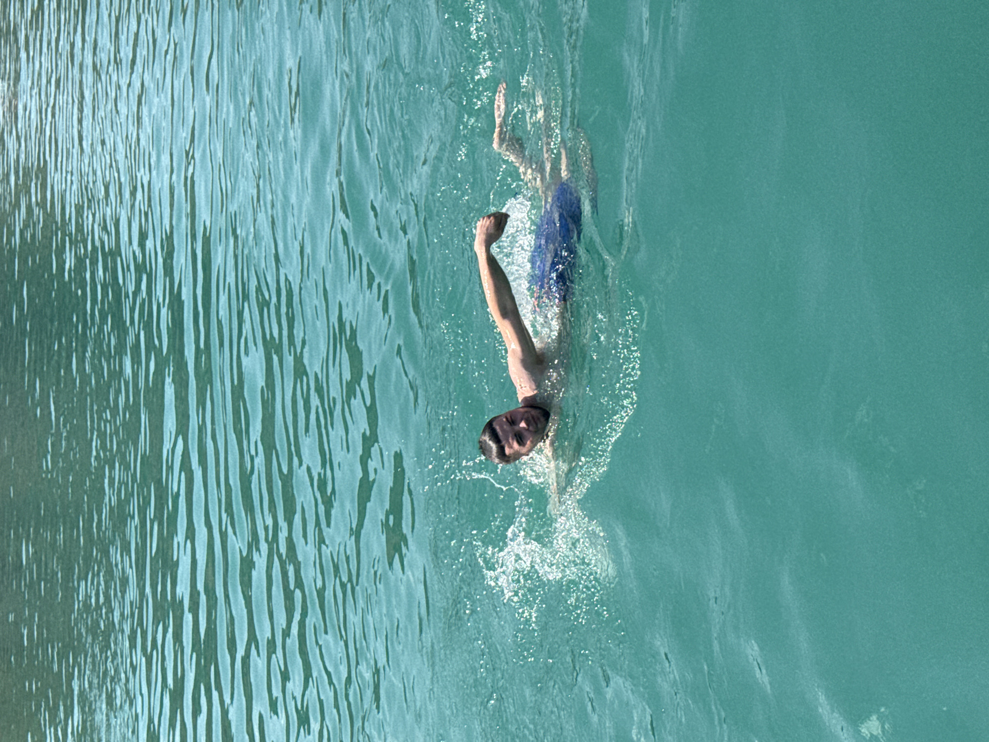 A male is swimming in the turquoise waters of the ocean. He is wearing blue swim trunks and appears to be in mid-stroke. The water is clear and calm with some ripples on the surface. The background is blurred, indicating the focus is on the swimmer. The location is the city of Karyer Izvestnyak, Russia.