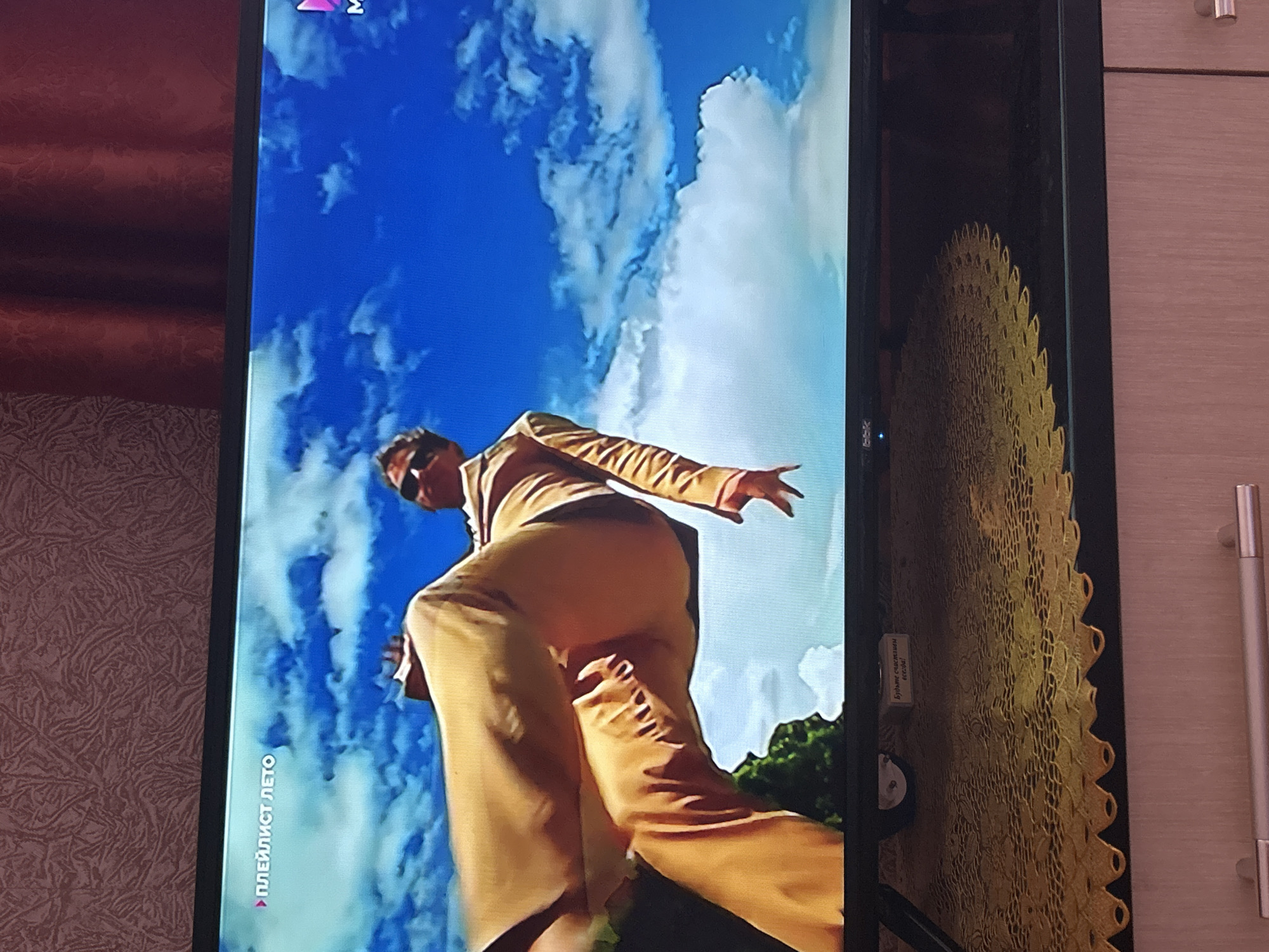 The image shows a television screen displaying a scene from a video. On the screen, a person is shown from a low angle, appearing to be in mid-air or levitating against a bright blue sky with white clouds. The person is wearing a light brown or tan suit with wide-legged pants and a jacket. They are also wearing sunglasses. The text "ПЛЕЙЛИСТ ЛЕТО" is visible in the top left corner of the screen, which translates to "Summer Playlist". On the bottom right of the screen, a pink "M" logo is visible, suggesting this is from a music channel or program. Below the television screen, there is a yellow decorative mat on a piece of furniture. A small white label with Russian text is visible on the bottom edge of the television screen; the visible text reads "Будьте счастливы". The scene is indoors, with a textured wallpaper and red curtains visible in the background, outside the frame of the television. There is no identifiable information in the image that indicates the location is Gelendzhik, Russia.