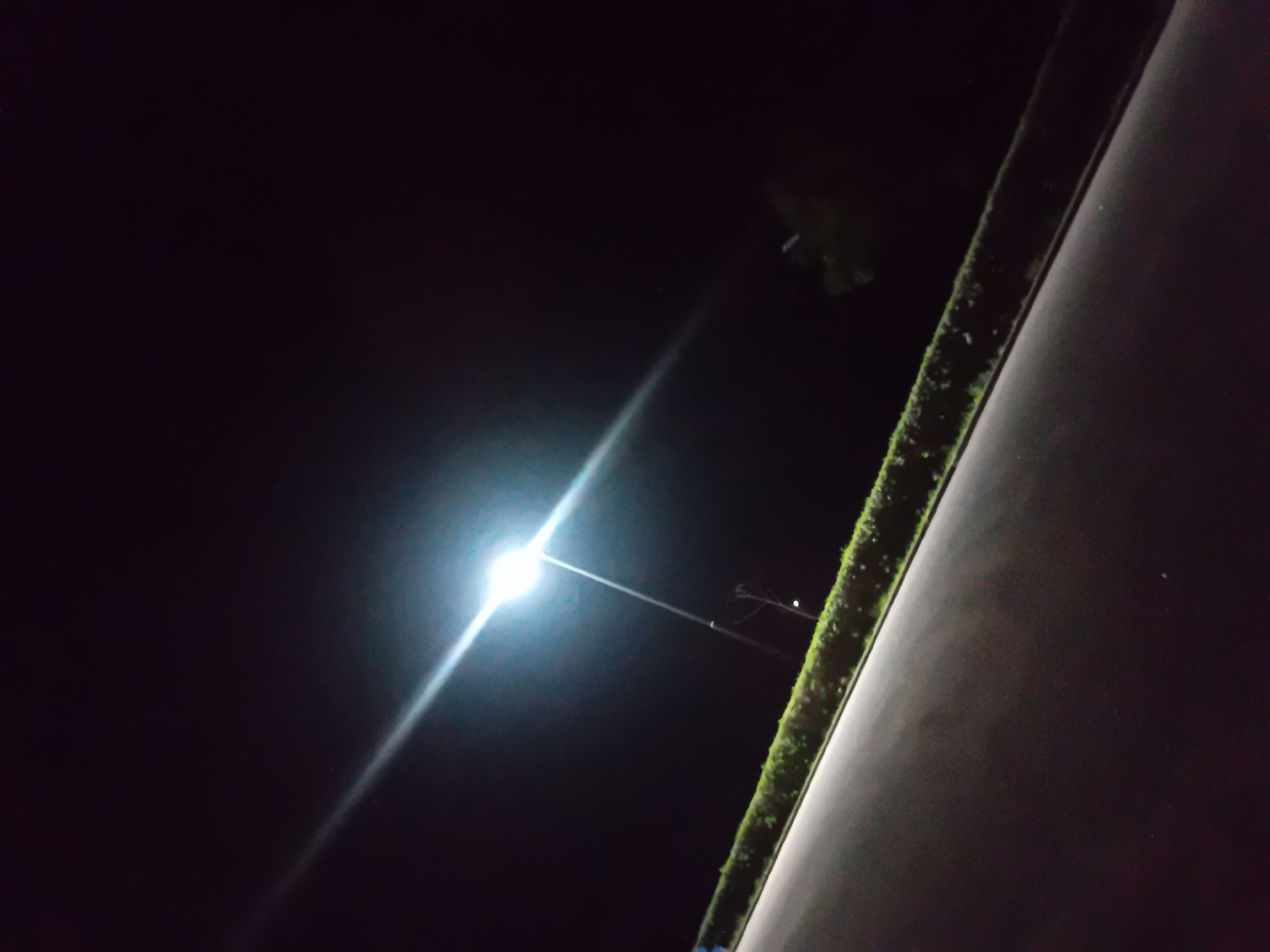 The image captures a dark, outdoor scene with a bright streetlamp illuminating a portion of the frame. A paved surface, likely a road or pathway, occupies the lower half of the image, angled upwards from left to right. Along the edge of this paved area is a line of dense, green vegetation, appearing as a manicured hedge.
Above the hedge, a tall streetlamp with a single bright light source dominates the upper left quadrant. The light from the lamp casts a strong glare and creates a visible beam that stretches across the dark sky. The sky itself is black, indicating nighttime, with only faint specks of light that could be stars.
In the background, behind the hedge and to the right of the streetlamp, there are dark shapes that suggest trees or other foliage, silhouetted against the night sky. A thin pole with wires is visible near the base of the streetlamp. The overall impression is that of a quiet, urban or suburban area at night, illuminated by artificial lighting. The location is unknown.