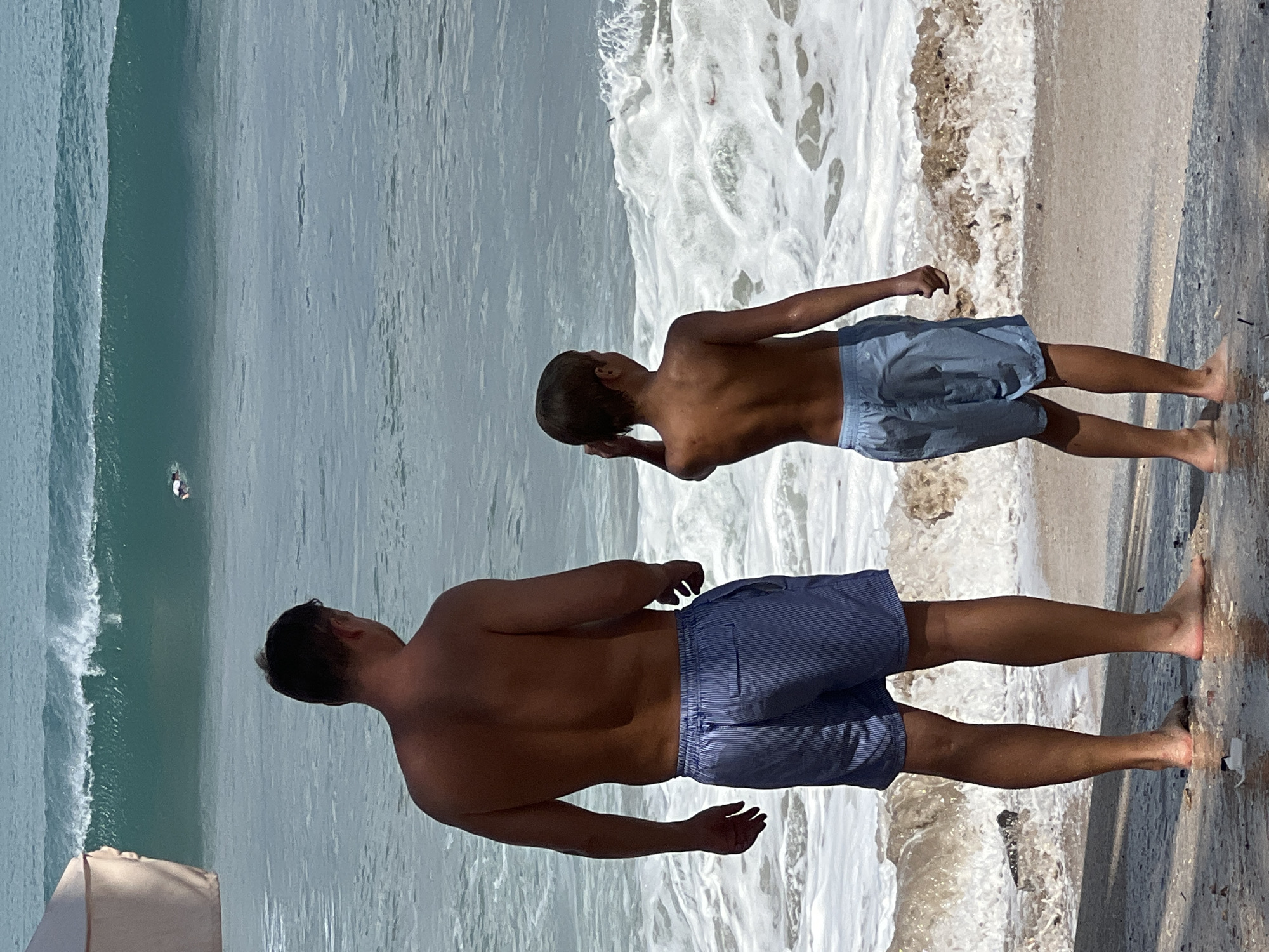 Two shirtless males, one adult and one child, stand on a sandy beach facing the ocean. The adult, on the left, wears blue and white striped shorts, and the child, on the right, wears light blue shorts. The ocean has large waves breaking, with a surfer visible in the distance. A light-colored beach umbrella is partially visible on the far left. The scene is likely Kuta, Indonesia, known for its surf.