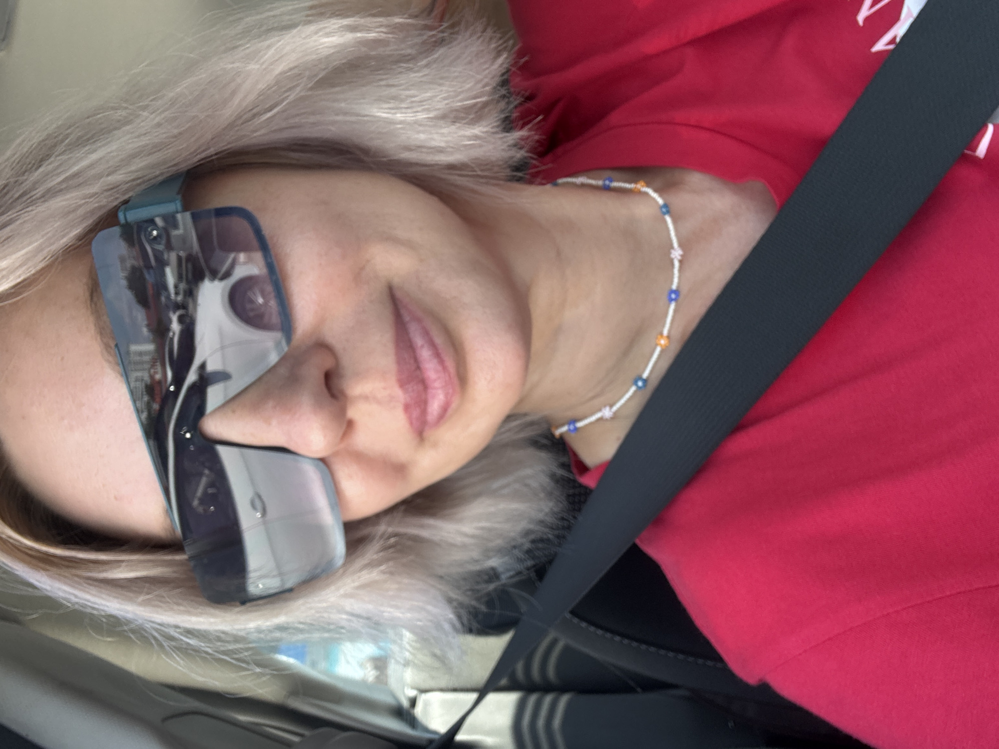 The image shows a close-up, waist-up shot of a woman wearing a bright red t-shirt and a beaded necklace. She has short, blonde hair and is wearing large, futuristic sunglasses with blue-tinted lenses that reflect cars and buildings. She is sitting in a car, as indicated by the seatbelt prominently crossing her chest and the visible car interior on the left side of the frame. The lighting suggests it is daytime and possibly sunny. There is no indication of the specific location being Sochi, Russia.
