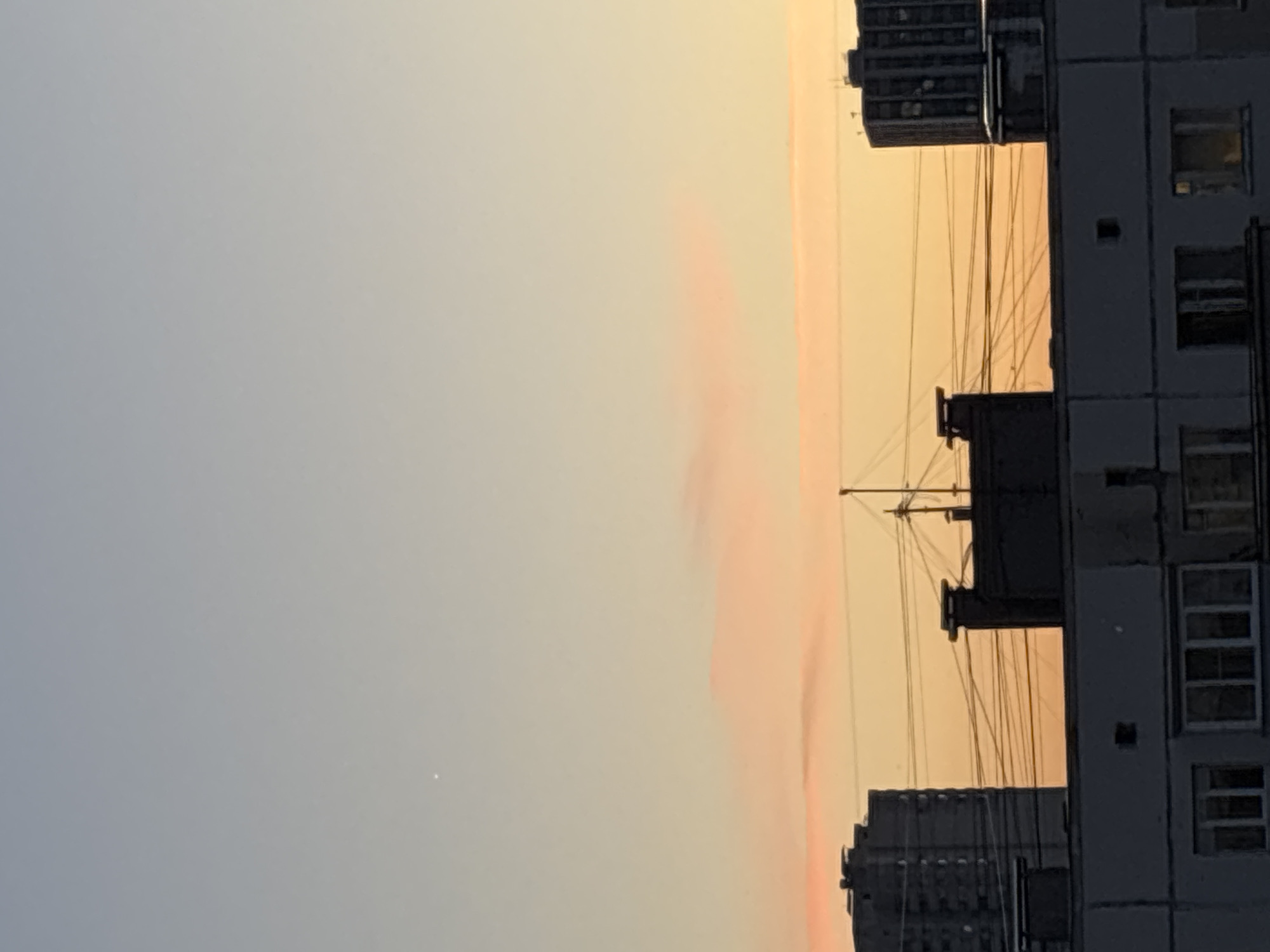 This is a photograph of a sky scene with buildings. The sky is a gradient of light blue to pale yellow and orange, with a few faint pink clouds visible. A single small, bright point of light, possibly a star or planet, is visible in the upper left quadrant of the sky. In the middle ground, there are several buildings. On the left is a tall, dark apartment building. In the center, there is a smaller, darker structure with a chimney and a pole with what appears to be an antenna or weather instrument. Further to the right is another tall apartment building. Numerous thin, dark wires crisscross the sky between the buildings. In the foreground, a portion of a lighter-colored, lower apartment building is visible, showing windows. The overall scene appears to be a view from a window or balcony in an urban environment, likely at dusk or dawn.