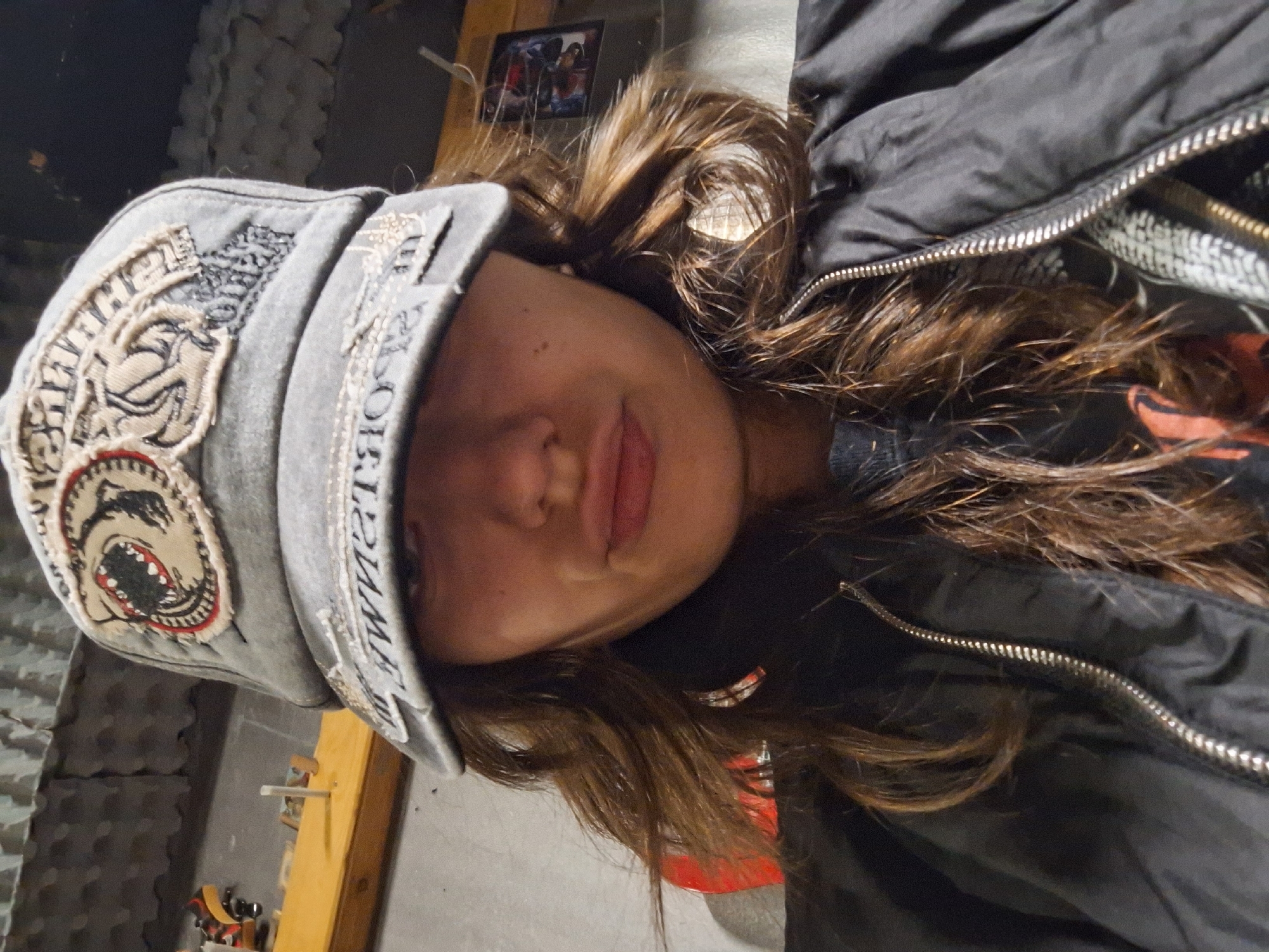 A young person is posing for a portrait. They are wearing a gray cap with decorative patches and text. The person has long, brown hair and is wearing a black jacket with a visible zipper. The background includes a textured gray wall and a wooden frame structure. A picture is mounted on the wall in the background. The location is not possible to determine from the image.