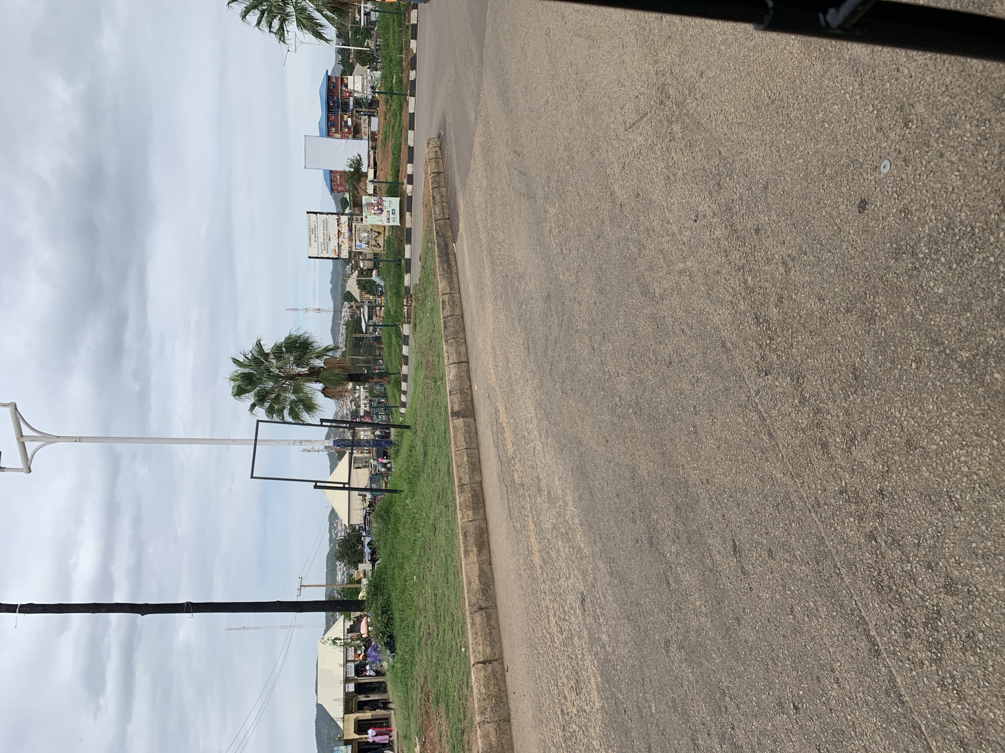 The image captures a wide-angle view of a street intersection or major thoroughfare in Rafin Par, Plateau State, Nigeria, under a uniformly overcast sky.
The foreground is dominated by a worn asphalt road surface, appearing light grey-brown with visible aggregate and minor cracks. This road is bordered on the left by a raised concrete curb, which separates it from a central grassy median. A small, circular white object, possibly a washer or disc, is visible on the road surface in the lower right quadrant.
The grassy median is unevenly maintained, with patches of greener and drier grass. Along its far edge, a black and white checkered curb or barrier is visible. Behind the median, the scene extends into a more developed area.
Prominently standing on or behind the median are several tall poles. To the far left, a slender, very tall dark black pole ascends towards the sky. Adjacent to it, a taller white pole with a T-shaped top, likely a street light pole, is positioned. A large, black, rectangular metal frame, presumably for a billboard, stands near the white pole. Further back and to the right, two large billboards are visible. One displays text, partially legible as "GOVERNMENT OF PLATEAU STATE" and "RACHEL PAR," with other smaller text. The adjacent billboard is blank white. Below these, a smaller poster with "GRACE" printed on it is visible.
To the far left of the image, a row of low-rise, single-story commercial buildings, some with awnings, are visible. Several pedestrians, including individuals in brightly colored clothing, are gathered or walking near these buildings, suggesting active street or market activity.
Beyond the median and billboards, additional structures include a distinctive two-story building with blue roofs and reddish-brown walls. A white-roofed building, possibly a public or religious structure, is situated to the left of the billboards. Indistinct vehicles, including what appears to be a tricycle (Keke Napep), and additional people are present in the mid-ground, but their specific actions or interactions are not clearly discernible due to distance.
Sparse palm trees are visible throughout the scene, including one prominent tree in the mid-right and others in the far distance. The horizon is marked by a line of low-lying hills or mountains under the cloudy sky, defining the geographic backdrop of the urban environment.