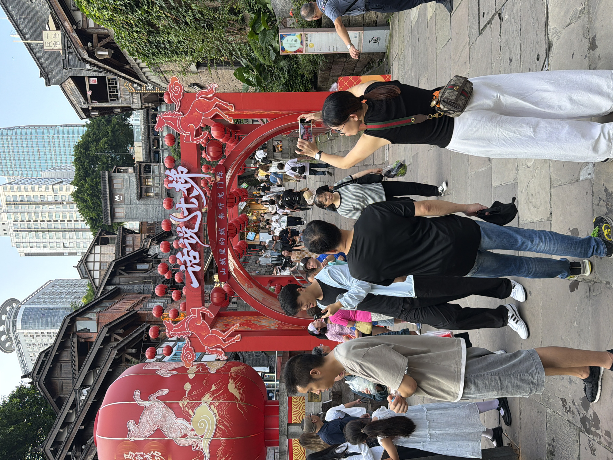This is a wide shot of a crowded pedestrian street in the Yuzhong District of China. The scene is dominated by a large, red, traditional Chinese archway adorned with horse motifs and lanterns. Behind the archway, traditional wooden buildings with tiled roofs are visible, stacked on a hillside. Modern skyscrapers rise in the background. A large red lantern is prominently displayed to the left of the archway.
In the foreground, a group of people are walking or standing. A woman in the center right of the frame is taking a photo with her phone, held horizontally. She is wearing a black tank top and white wide-leg pants, with a patterned crossbody bag. To her left, a man in a black t-shirt and jeans is walking away from the camera. To the left of the woman, a young man in a light blue jacket and black t-shirt is holding a phone. In front of him, a man in a grey t-shirt and shorts is looking to the left. Other individuals are visible throughout the scene, including a young girl in a white dress on the far left. The street is paved with stone. The overall atmosphere appears to be festive and busy.