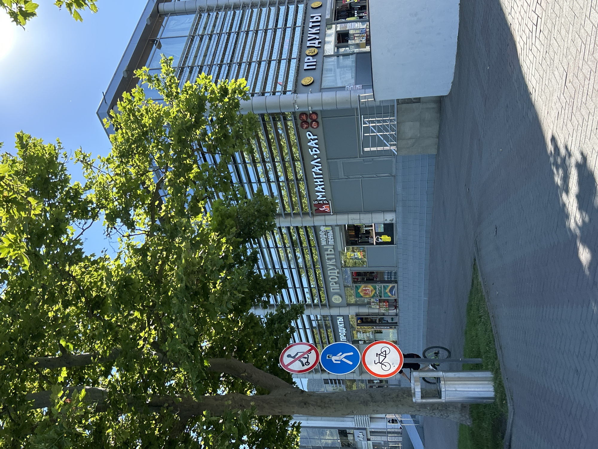 The image shows a modern building with a glass facade on a sunny day. In front of the building, there is a tree and a paved sidewalk. Several traffic signs are visible on a pole next to the tree. One sign depicts a person on a scooter with a red circle and a slash, indicating "no scooters." Below that, a blue sign shows a person walking, signifying a pedestrian zone. The bottom sign shows a bicycle, indicating a bicycle path.
The building houses several businesses. One sign reads "ПРОДУКТЫ" (Products) with a clock symbol, suggesting it is open 24 hours. Another sign, "МАНГАЛ-БАР" (Mangal-Bar), indicates a grill bar. The building also has the "ПРОДУКТЫ" sign again on its right side. A person on a bicycle is visible riding past the signs and the tree. A metal trash can is located next to the tree. The pavement is made of interlocking bricks. The context of Gelendzhik, Russia, suggests a coastal city environment, though this specific view focuses on the urban streetscape.
