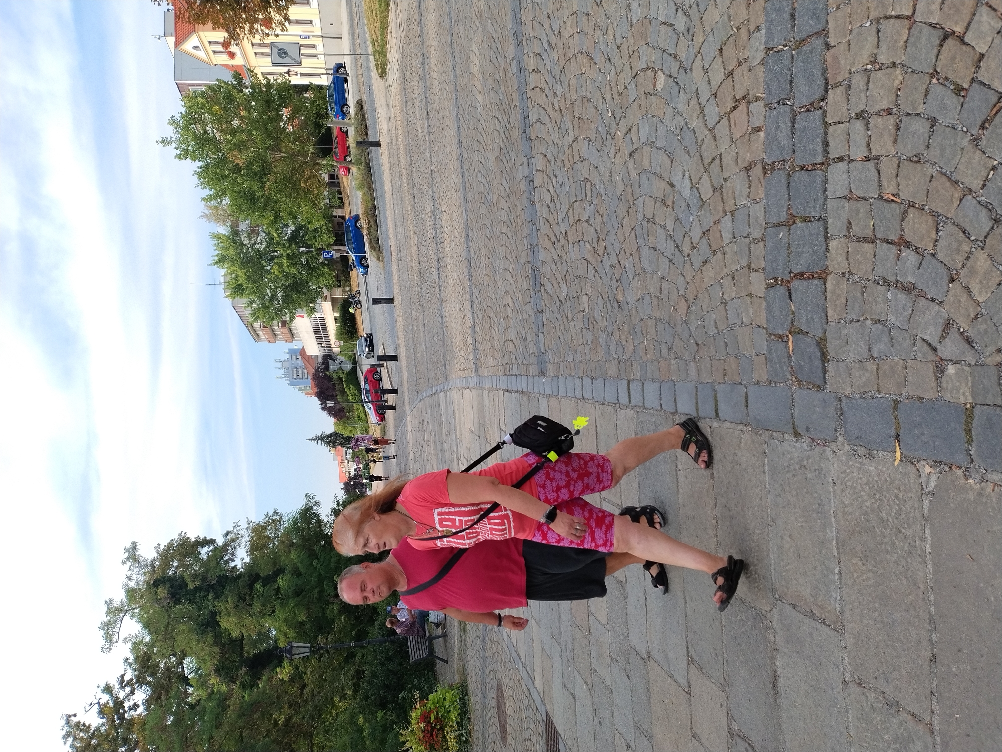 The image depicts a paved walkway with cobblestone sections and large stone slabs, characteristic of the area around the Písek Bridge in Písek, Czechia. In the foreground, a couple walks along the path. The man, wearing a red t-shirt and dark shorts, walks with his left hand by his side. To his right, a woman with blonde hair, wearing a pink t-shirt and patterned shorts, carries a black crossbody bag. Behind them, on a bench to the left, two individuals are seated. The background shows trees, buildings, and a street with several parked cars, including a blue sedan and a red hatchback, suggesting an urban environment. The sky is partly cloudy.