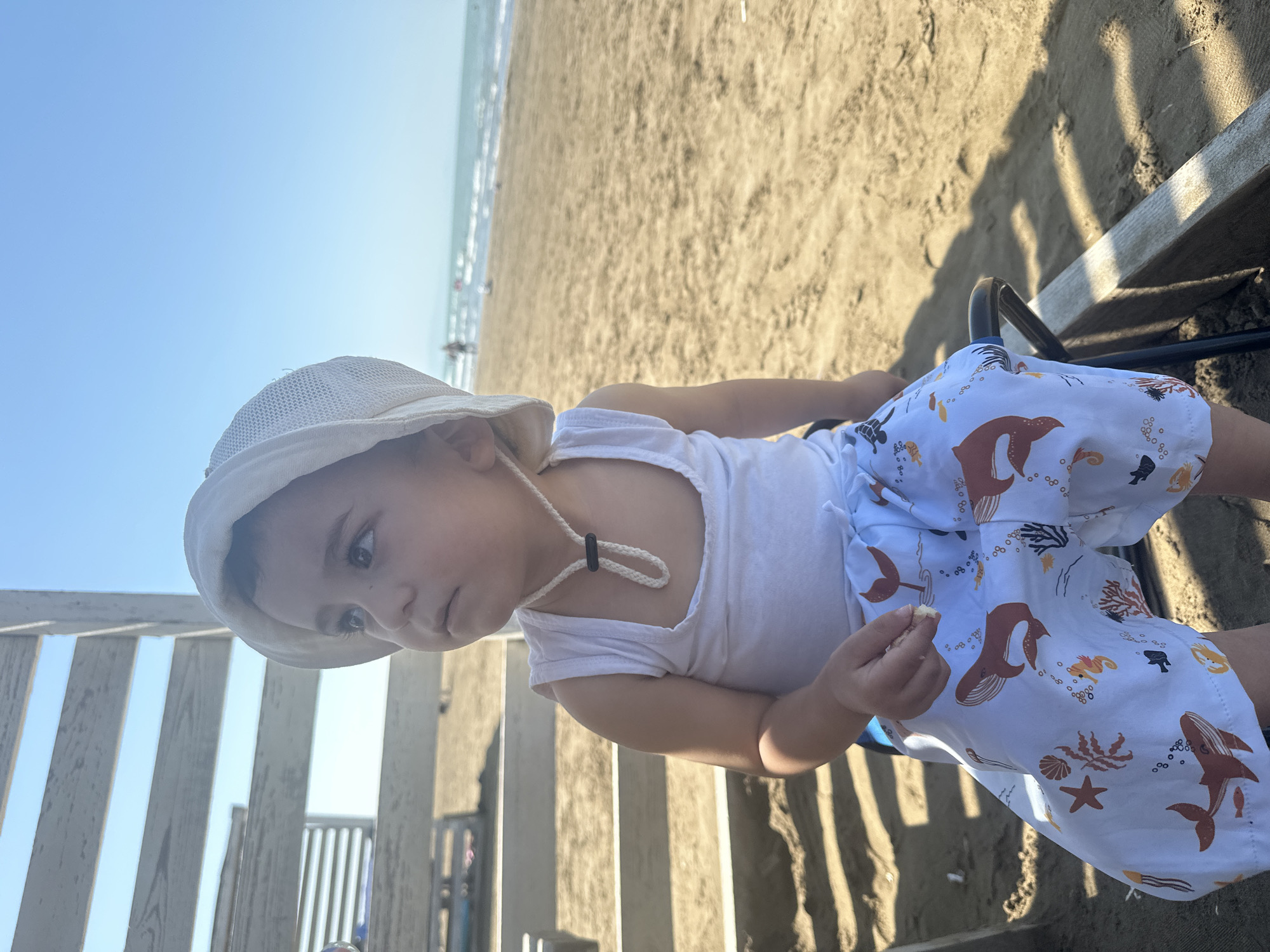 A young child wearing a white hat and a white tank top with patterned shorts sits on a chair on a sandy beach. The child is looking to the left, with a piece of food in their right hand. In the background, the ocean and a clear blue sky are visible, with some distant figures walking along the shoreline. Wooden structures are present to the left of the child. The scene appears to be in the city of Sabunchu Raion, Azerbaijan, likely a public beach.