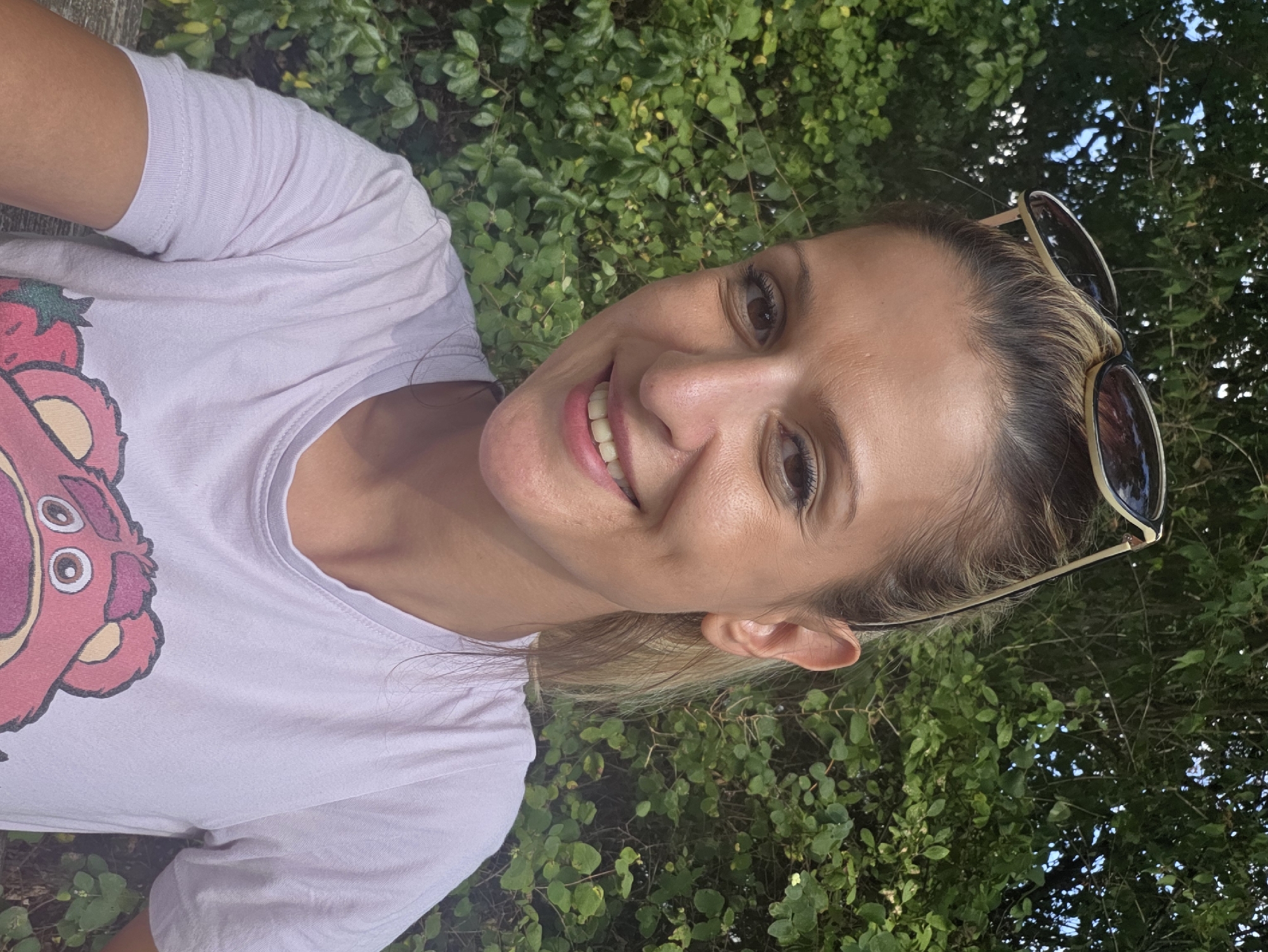 A woman with blonde hair pulled back, wearing sunglasses on top of her head and a light purple t-shirt with a cartoon bear graphic, smiles at the camera. She is outdoors, with a dense green hedge as the background. The image appears to be a selfie taken in a park or natural setting.