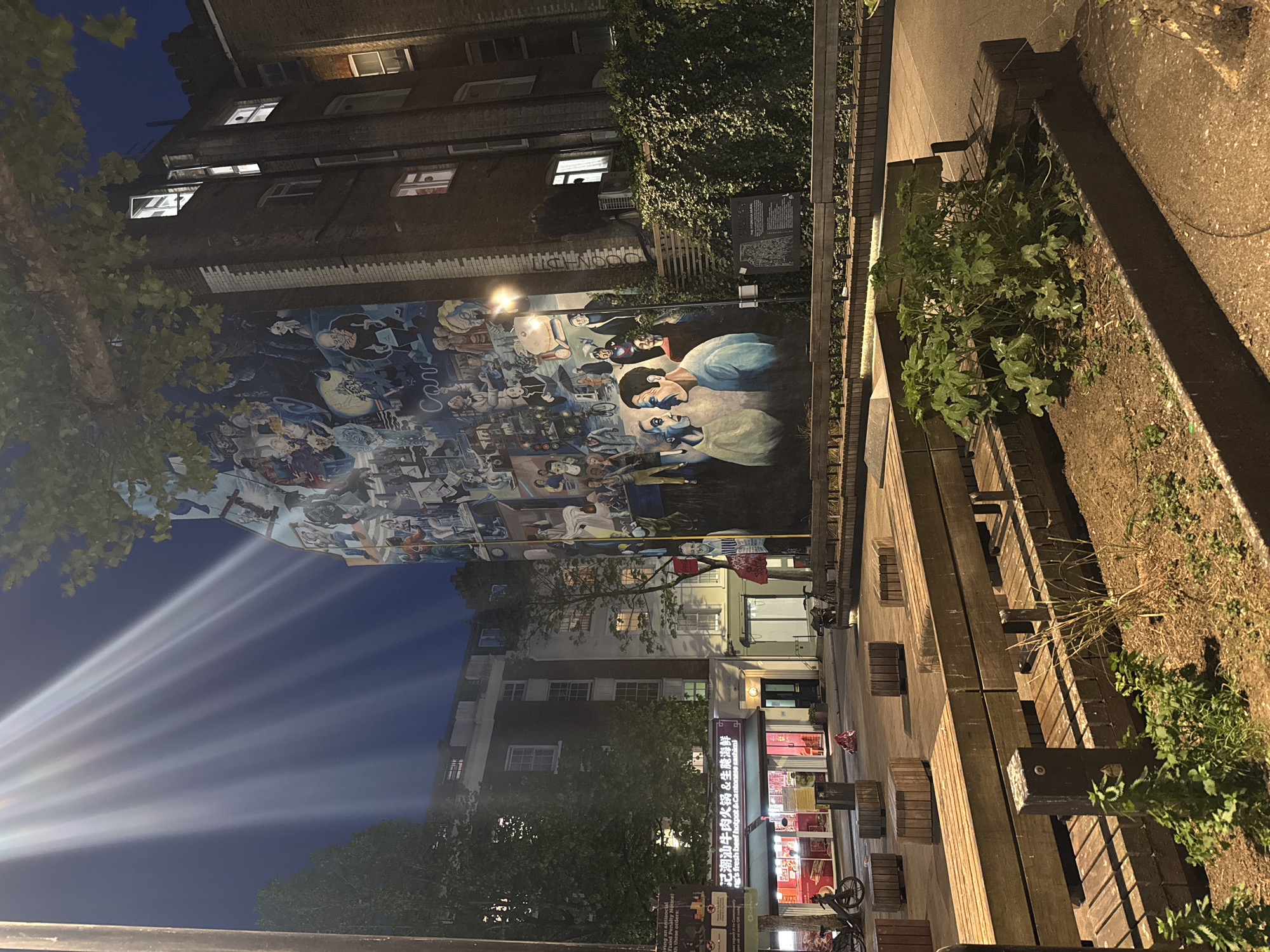 This night-time image captures a bustling urban scene, likely a pedestrian street or courtyard in London, United Kingdom. The shot is vertically oriented, showing the tops of buildings reaching into a dark blue sky.
A striking, multi-panel mural dominates a large wall on the left, depicting numerous stylized figures and scenes in vibrant colors, suggesting a rich narrative or artistic expression. Above the mural, the building is made of brick, with several windows, some illuminated from within, casting a warm glow. Trees with green foliage are visible in the foreground and beside the buildings, adding a touch of nature to the concrete environment.
To the right, a series of wooden benches and planters creates a public seating area, illuminated by warm streetlights that cast long shadows. Lush green ivy climbs a nearby wall, where a dark informational plaque is mounted. Further down the street, more buildings with storefronts are visible. One prominent shop signage displays Chinese characters "记潮汕牛肉火锅&生腌海鲜" (Chaoshan Beef Hot Pot & Raw Seafood), with an English translation below: "g's fresh beef hotpot & Cantonese sashimi". Bicycles are parked near the storefronts.
A powerful light source, possibly a streetlight or lamp, creates dramatic, radiating beams across the dark night sky in the lower left portion of the image. Other visible text includes "Car" potentially on the mural or as graffiti, and fragmented letters like "Pena", "AT", "A", and "B" from various signs or markings. The overall atmosphere is quiet and contemplative, highlighting the artistry of the mural against the nocturnal city backdrop.