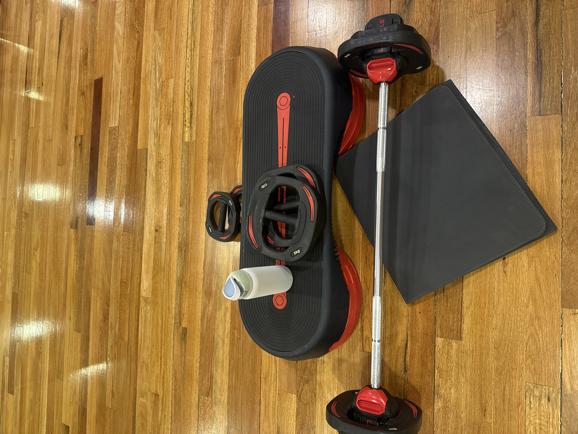 The image shows a collection of fitness equipment arranged on a wooden floor. The floor is composed of light-colored wooden planks, with variations in the grain and coloration. The equipment includes a black and red workout platform with a curved shape, several weight plates (marked with "5kg" and "1.1lb"), a barbell with weights attached, and a dark gray rectangular mat. A white water bottle is placed on the platform. The lighting appears to be indoor, with a slightly warm tone. There is no information in the image to indicate its location in Puebla, Mexico.