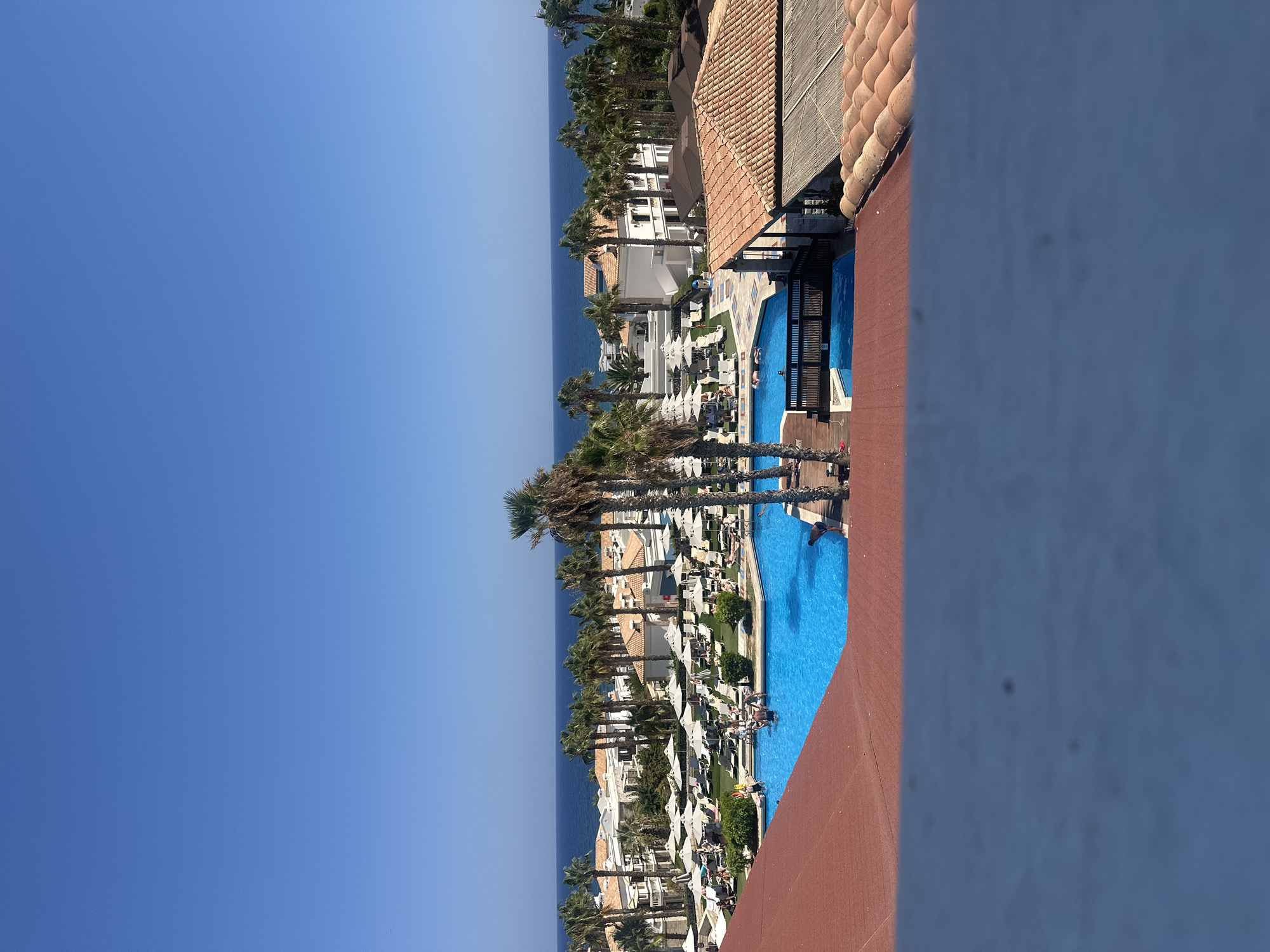 The image captures a vibrant poolside scene in Chersonisos Municipal Unit, Greece, under a clear blue sky. In the foreground, a red-tiled roof and a blurred blue wall suggest a vantage point from a building. The main focus is a large, brightly colored blue swimming pool filled with water. Several people are visible in and around the pool; some are swimming, while others are relaxing on lounge chairs beneath white umbrellas scattered across a green, manicured lawn.
Palm trees are a prominent feature, lining the sides of the pool area and extending towards the background. Beyond the pool and palm trees, a complex of white buildings with terracotta roofs is visible, hinting at a resort or hotel complex. Further in the distance, the deep blue expanse of the sea meets the horizon, with a faint line of coastline visible. The overall impression is one of a sunny, idyllic vacation destination.