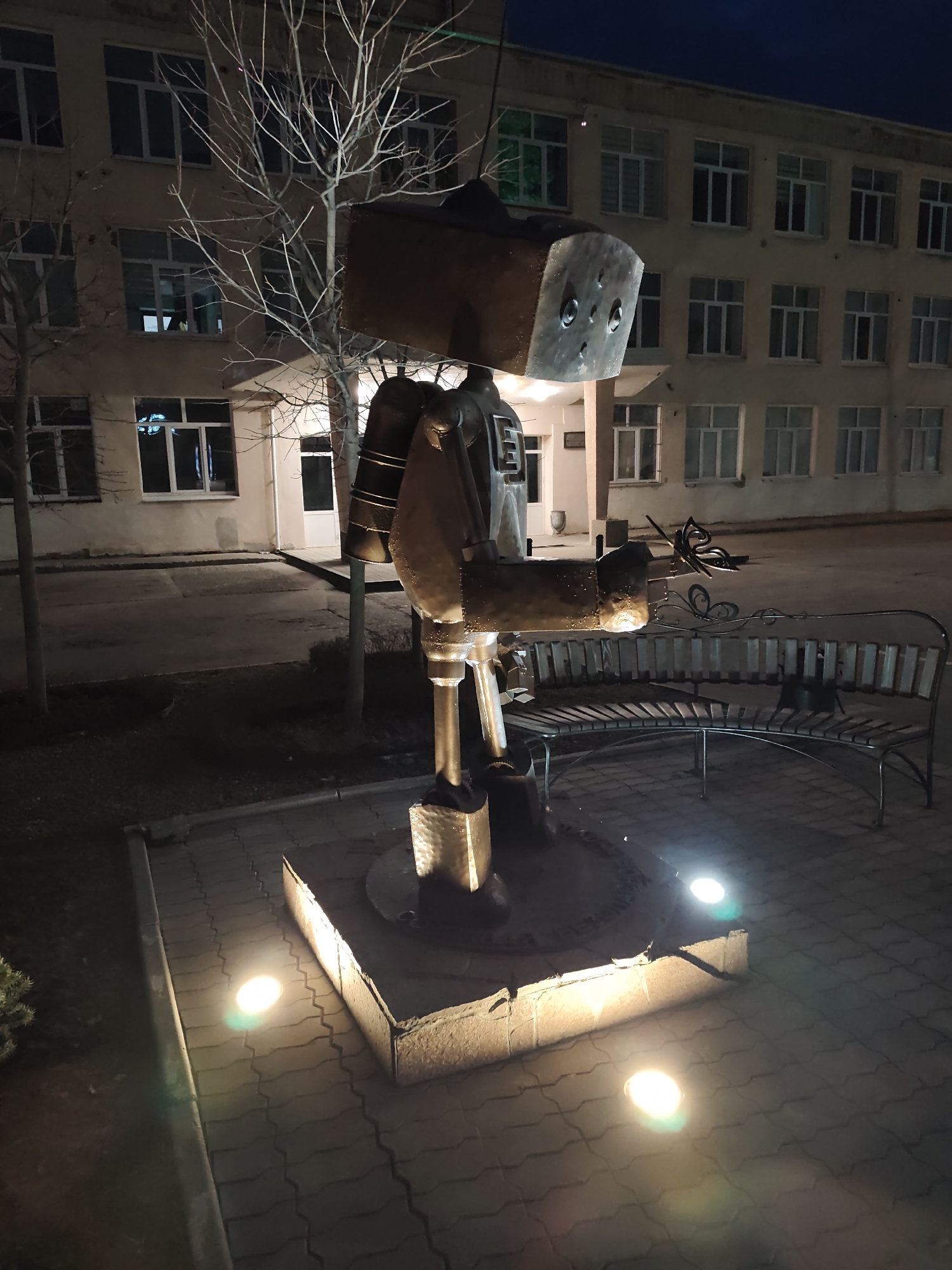 The media file captures an outdoor scene at night in Chaikovsky, Russia, dominated by a distinctive metal sculpture. The central subject is a large, stylized figure, possibly a robot or character, crafted from various metallic components. It features a blocky, rectangular head with two prominent circular "eyes," a robust body, and an arm extending forward, holding what appears to be an ornate metal butterfly. The sculpture stands on a raised concrete base, illuminated by two ground-level spotlights, one emitting a warm yellow glow and the other a cooler blue light, casting reflections on the surrounding grey interlocking paving stones.
To the right of the sculpture, a curved wooden bench with metal supports is visible, potentially part of the same artistic installation, as a decorative metal butterfly ornament is attached near its backrest. Several bare-branched deciduous trees stand behind the sculpture and bench, suggesting a late autumn or winter setting. In the background, a multi-story building with numerous evenly spaced windows serves as the backdrop. The building appears to be light-colored with dark window frames, and lights are visible in some of the interior windows. The sky above is a deep, dark blue, indicative of nighttime. No people are visible, and no discernible text is present in the image. The scene is static, highlighting the public art installation under artificial illumination.