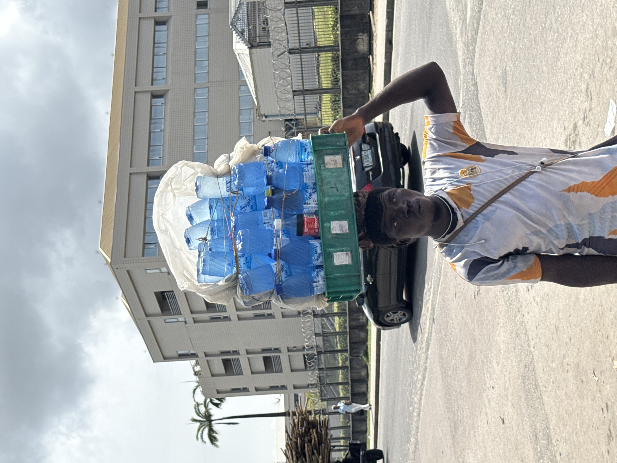 A young man carries a large stack of bottled water on his head, standing on a paved street in an urban environment. He is positioned slightly off-center to the right, looking directly at the viewer with a neutral expression. He has dark skin and wears a light-colored, short-sleeved shirt with an abstract pattern of orange and dark grey/black shapes. A thin strap, possibly from a bag, crosses his left shoulder.
The impressive load on his head consists of multiple layers of blue plastic water bottles, some clear bottles, and a red can labeled "SPRITE" visible within a green plastic crate. A white plastic bag is draped over the top of the bottles. His right arm is raised, bent at the elbow, seemingly to help with balance.
In the background, a modern, multi-story building with numerous vertical windows stands prominently. A dark-colored car is parked behind the man, with its license plate "KJA 241" partially visible. A fence topped with barbed wire separates the street from a green area, and a few palm trees are visible further in the distance. The sky is bright with scattered clouds, indicating it is daytime. The scene captures a street vendor in a bustling urban area, consistent with the provided location of Lekki, Nigeria.