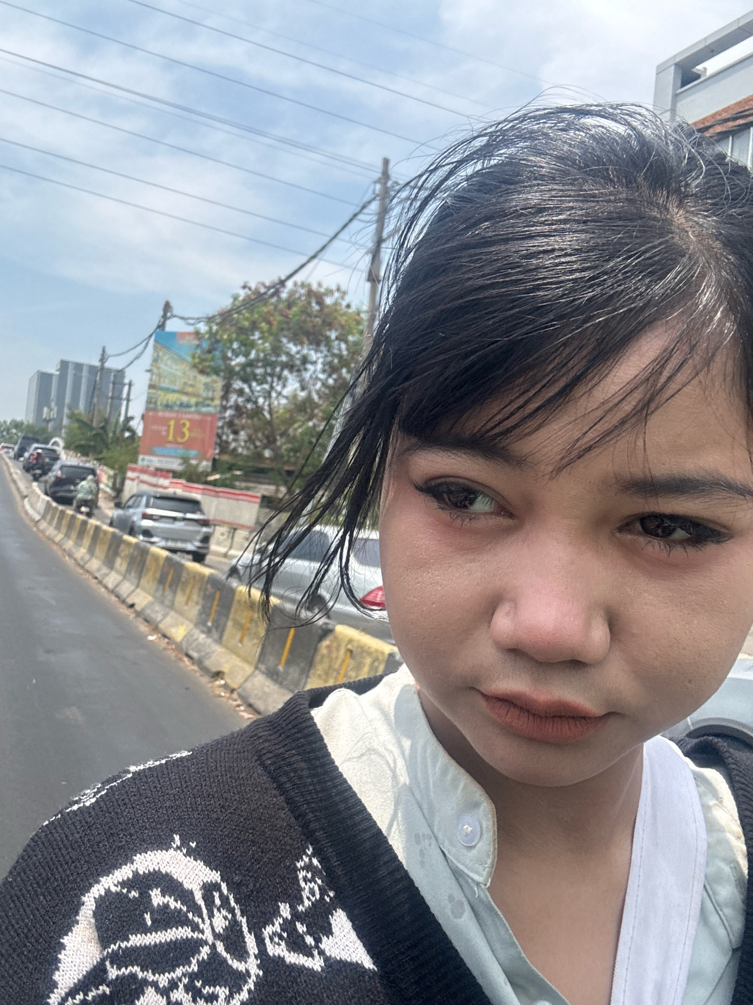 In a medium-angle, close-up selfie taken outdoors, a young woman with dark hair stands near a street. She is wearing a black and white patterned cardigan over a light blue collared shirt. Her facial features are prominent, with her dark eyes looking to the side. The background reveals a street lined with cars and concrete barriers, indicating an urban environment. Tall buildings and trees are visible in the distance, along with power lines and a billboard with the number "13" on it. The scene appears to be a roadside in North Jakarta, Indonesia, under a bright, cloudy sky.