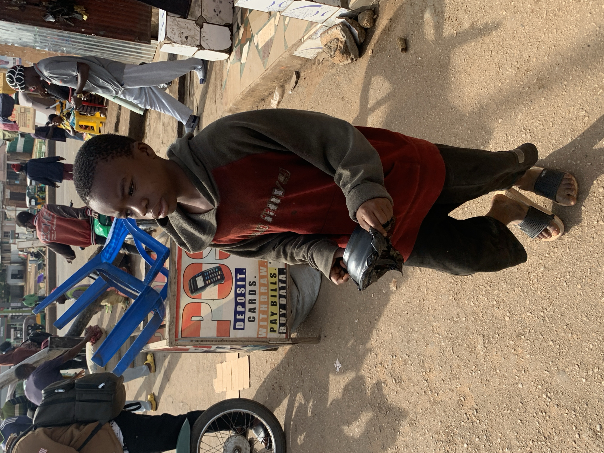 A young boy, likely of Nigerian descent, stands in an outdoor setting, possibly a street or market in Gura Top, Nigeria. He is wearing a red and grey hooded sweatshirt and dark pants, with dark-colored sandals on his feet. In his hands, he holds a dark, bowl-like object wrapped in black plastic.
Behind the boy, a wooden stall with brightly colored signage is visible. The sign includes text such as "DEPOSIT," "CARDS," "WITHDRAW," "PAY BILLS," and "BUY DATA," suggesting it is a service point for financial transactions or mobile services. Stacked blue plastic chairs are placed in front of the stall.
In the background, other individuals and elements of a street scene are present, including people walking, a motorcycle wheel, and various structures, all under a bright, possibly daytime, sky. The ground is a mix of dirt and gravel.