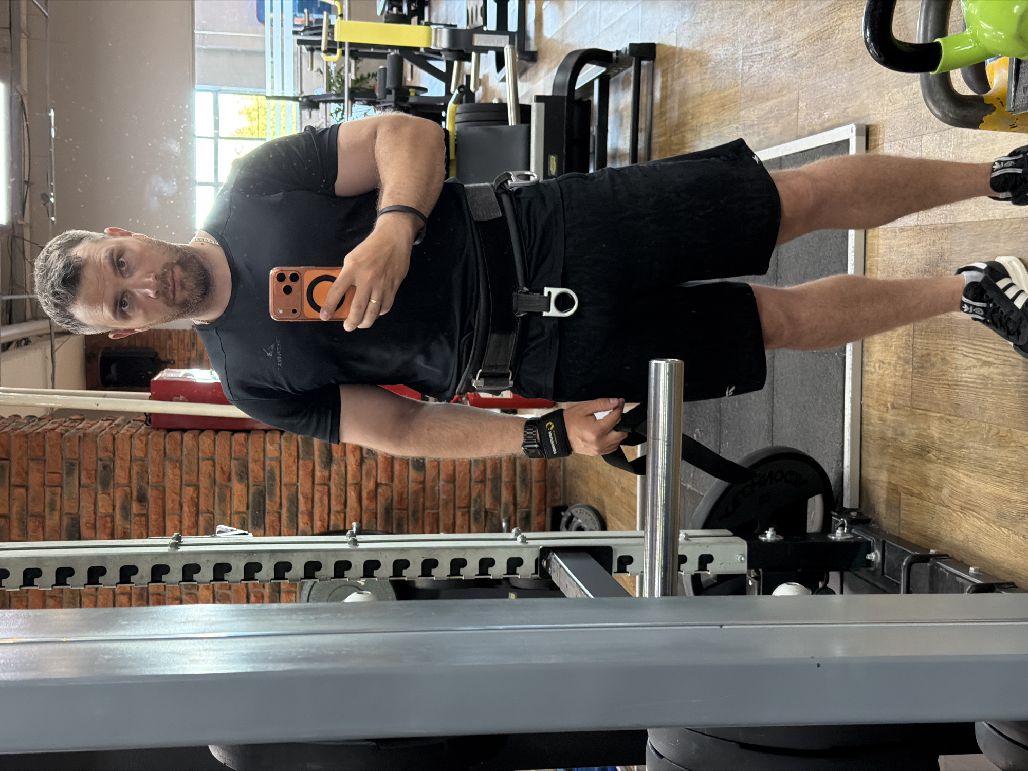 The image shows a man in a gym setting. He is dressed in a black t-shirt and black shorts, and he is wearing a weightlifting belt. He is holding a phone in his right hand, likely taking a selfie. The gym equipment in the background includes a leverage squat machine, weights, and a cable machine. The background also shows a red brick wall and a window. There is a reflection of the gym in a mirror. The location is potentially in Chaikovsky, Russia, as per the prompt.