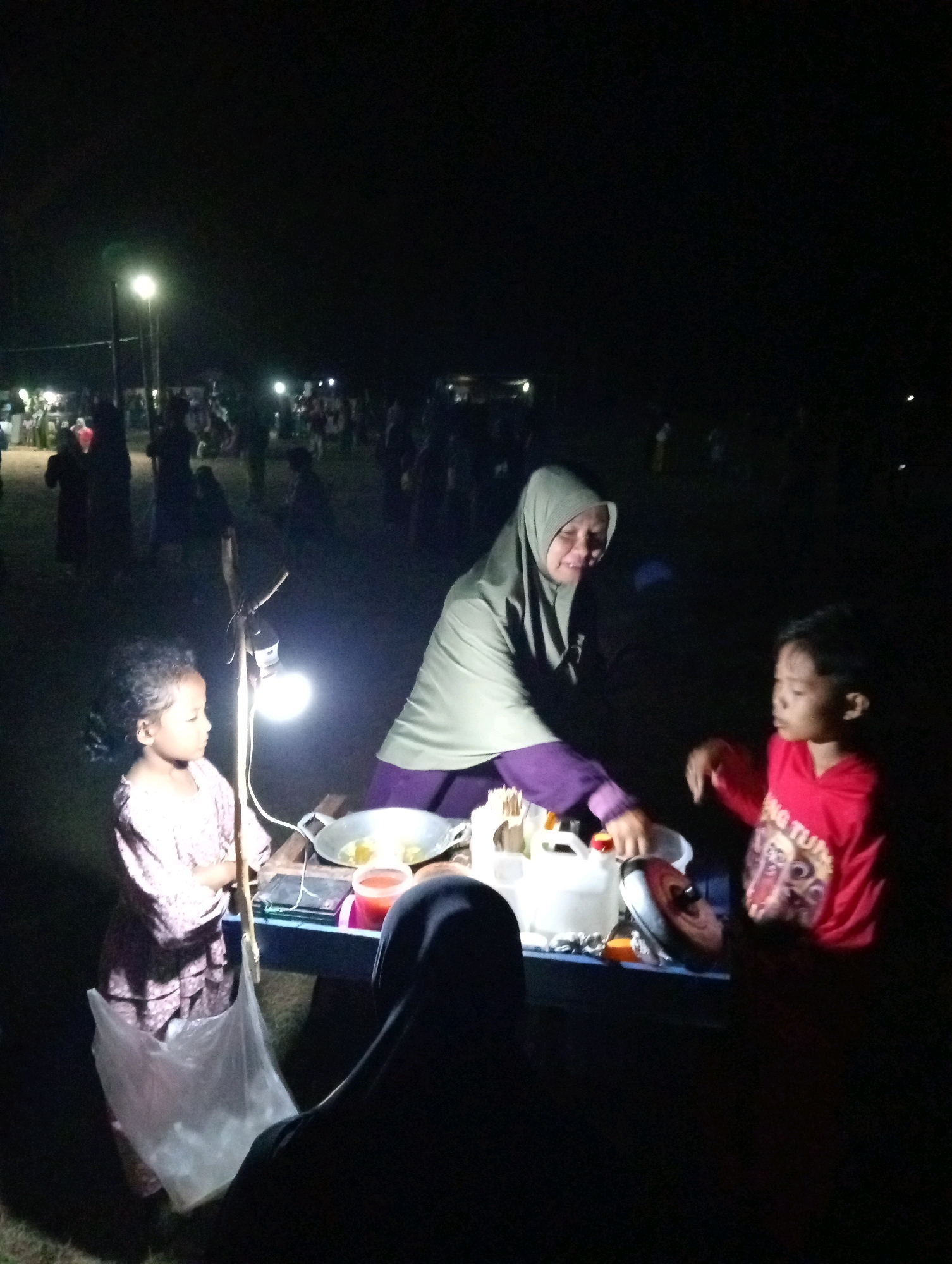 In the dimly lit environment of Pagerungan Kecil, Indonesia, a nighttime scene unfolds with several individuals gathered around a makeshift food stall. The primary focus is on a woman wearing a hijab and a purple dress, who appears to be serving or preparing food. She is standing behind a blue table laden with various items, including cooking utensils like a wok and a white container, as well as what looks like a bottle of red sauce.
To her left, a young girl with curly hair, dressed in a pink floral patterned garment, stands attentively. She is holding a clear plastic bag filled with items. To the woman's right, a young boy in a bright red shirt is positioned, seemingly interacting with the scene. Partially visible in the foreground, a person wearing a black hijab is seated, their back to the camera, observing the activity.
The scene is illuminated by a single, bright light bulb hanging from a makeshift stand, casting a harsh glow on the immediate surroundings and creating deep shadows. In the background, blurred figures and lights suggest the presence of other people and possibly more stalls or activities, indicative of a communal gathering or market at night. The ground appears to be covered in grass. The overall atmosphere is that of an outdoor evening gathering in a rural or semi-urban Indonesian setting.