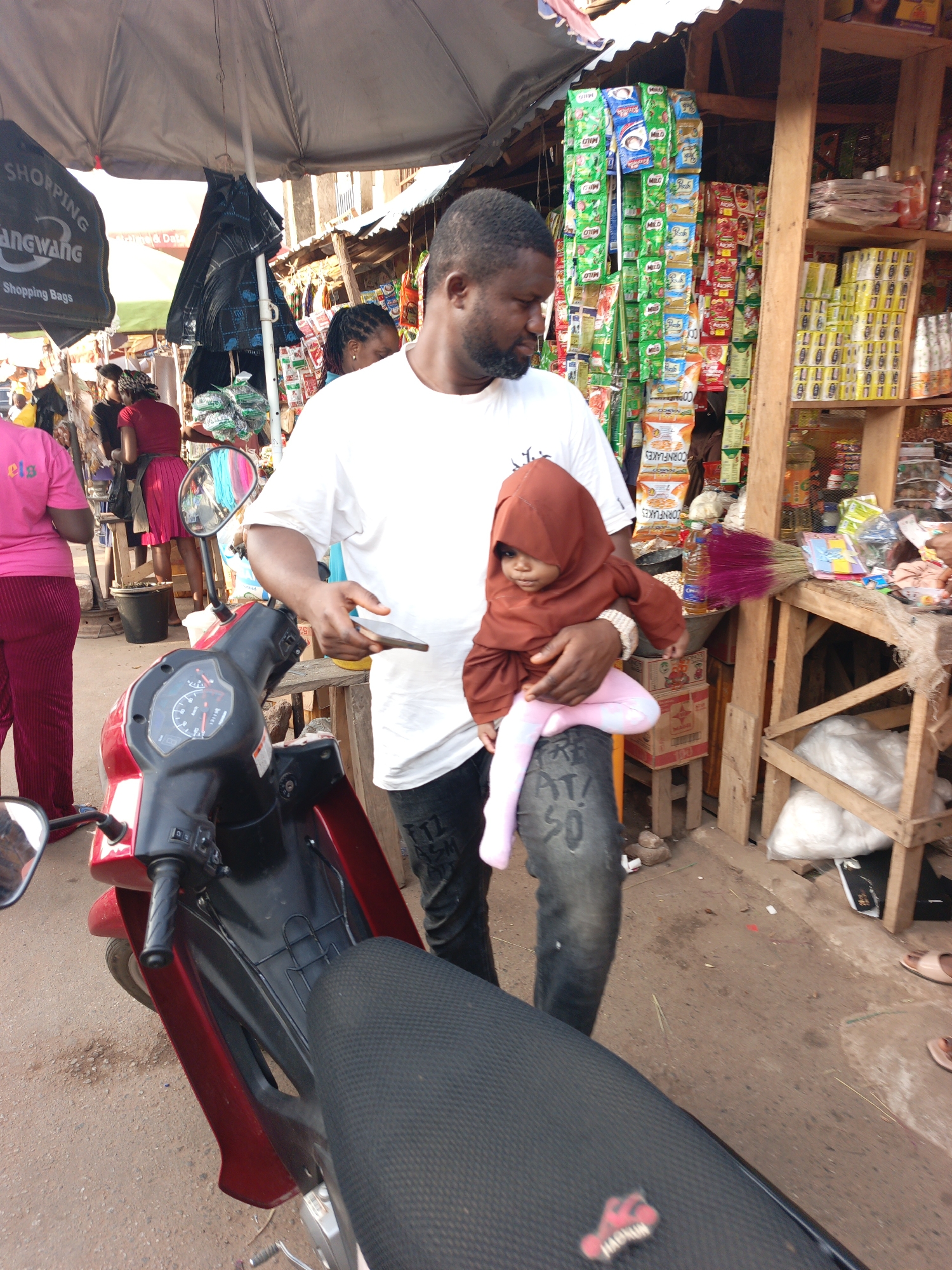 In an outdoor market in Akwanga, Nigeria, a man stands next to a red and black motorcycle, holding a baby in a brown hooded outfit and pink leggings. The man is holding a cell phone. Behind him, a wooden stall is filled with packaged goods, including Milo, cornflakes, and other food items. A large umbrella provides shade to the man and child. Other people are visible in the background, including a woman in a pink shirt and red pants. The ground is dirt.
