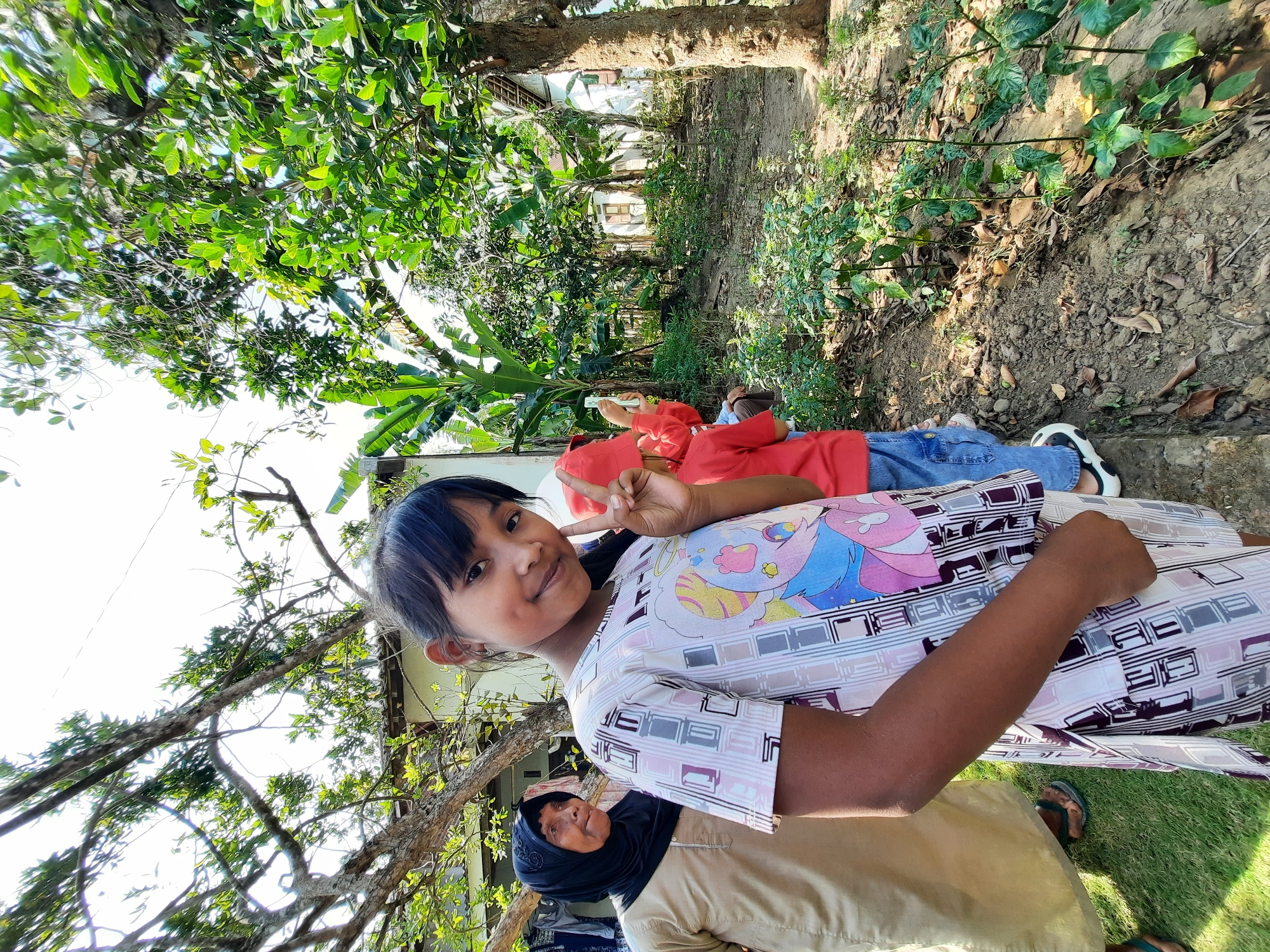 In Lugu, Indonesia, a young girl with dark hair and a light-colored t-shirt featuring a cartoon character and geometric patterns stands in the foreground, making a peace sign with her right hand. She has a dimple on her left cheek. Behind her to the left, an older woman wearing a beige garment and a black hijab is partially visible. Further back, two children in red shirts and blue jeans are observed, with one holding up a mobile phone. The environment is a garden with lush green trees, including banana trees, and a rustic house in the background. The ground is covered with dry leaves and soil, suggesting an outdoor, possibly rural, setting. The lighting indicates it is daytime with bright sunlight filtering through the trees.