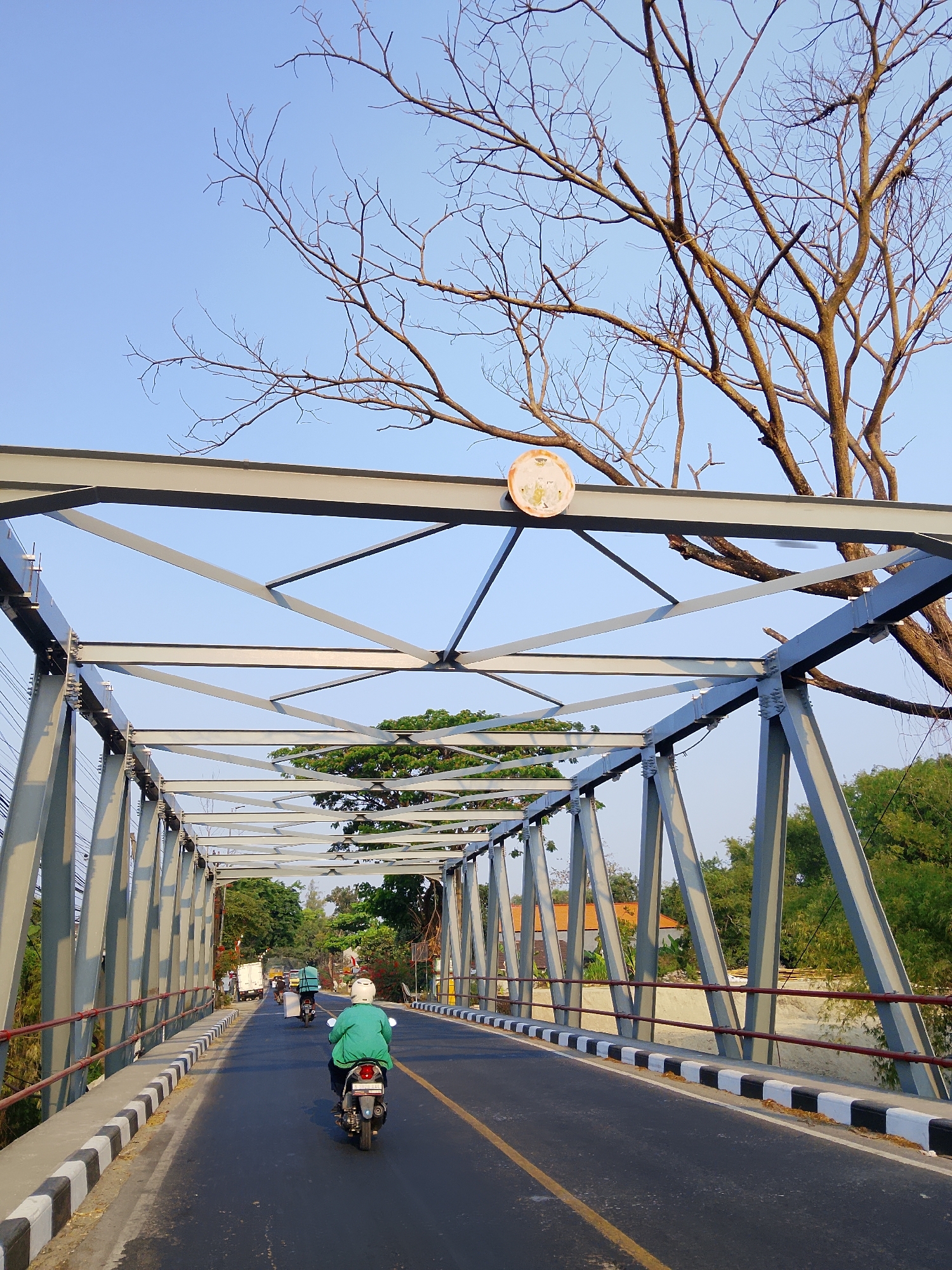 An outdoor, eye-level, full shot captures a view of a truss bridge on a sunny day. The sky is a clear, light blue, with the bare branches of a tree reaching into the frame from the top right. The bridge's grey metal structure dominates the foreground and midground. The road surface is asphalt with a yellow dividing line and a black and white striped curb. Two motorcyclists are visible on the bridge, both traveling in the same direction away from the viewer. The lead motorcyclist wears a green jacket and a white helmet, while the second motorcyclist also wears a white helmet and a lighter colored jacket. In the distance, there is a white truck and other vehicles. Lush green trees and some orange-roofed buildings are visible beyond the bridge on either side. A circular sign with an emblem is attached to the top of the bridge structure. The scene appears to be on a road in the city of Banjarjo, Indonesia, likely an area with a rural or semi-rural setting given the vegetation.