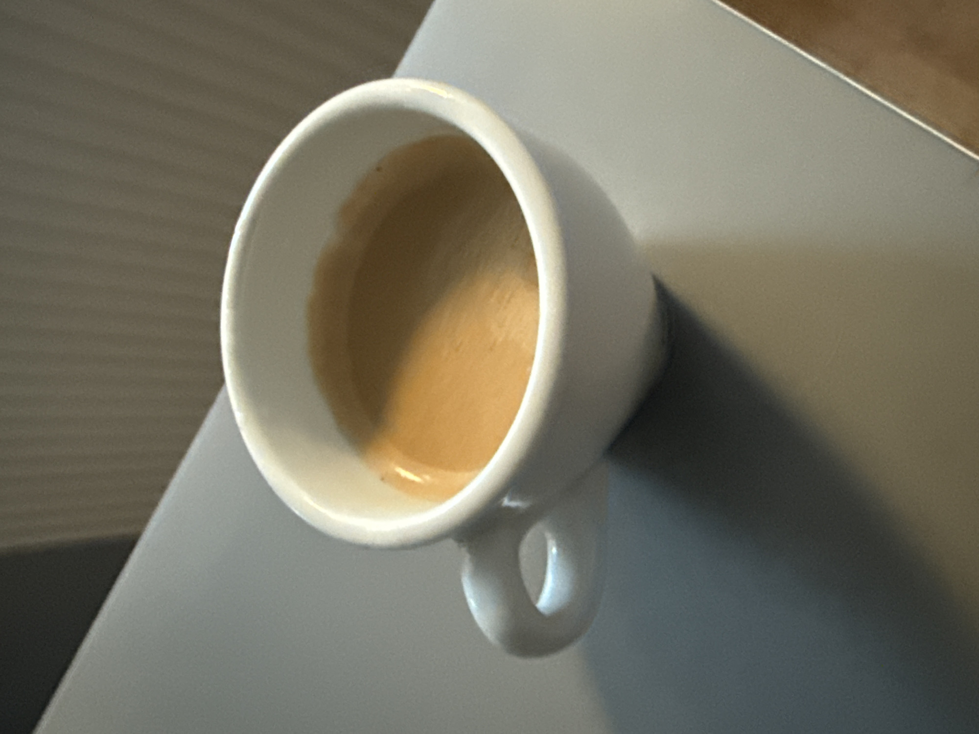 A close-up, eye-level shot features a white ceramic espresso cup filled with coffee. The coffee has a light brown crema and appears to be freshly brewed. The cup has a small, C-shaped handle. The cup rests on a smooth, grey surface, possibly a table or countertop. In the background, a vertically ribbed texture is visible, suggesting a wall or radiator cover. The lighting creates a shadow from the cup onto the grey surface. The image does not provide any information that would allow for its location to be identified in Wrocław, Poland.